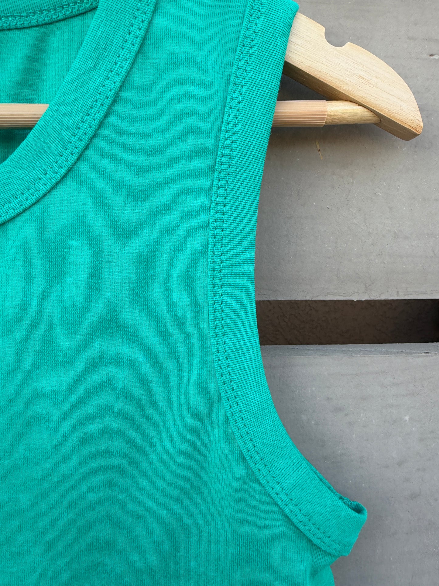 Close-up of a bright teal sleeveless top hanging on a light wood hanger against a gray wooden surface.