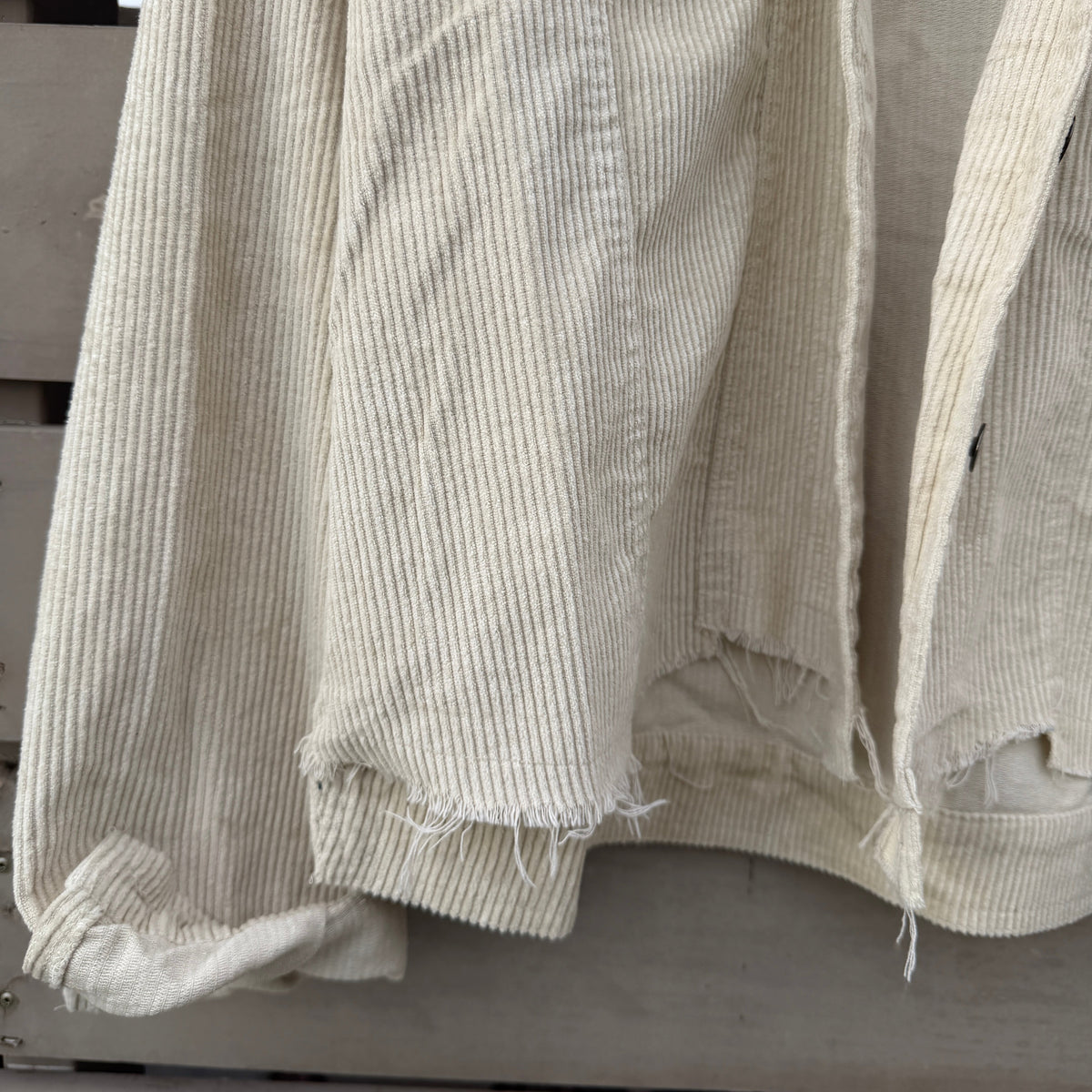 A close-up of a light beige ribbed corduroy jacket with raw, frayed hems, hanging against a weathered gray wooden background.