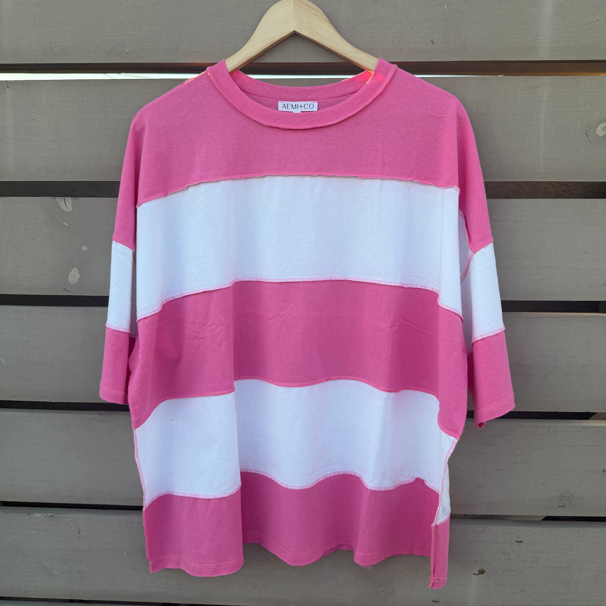 A pink and white horizontally striped short-sleeve t-shirt hanging on a wooden hanger against a gray wooden slat wall.