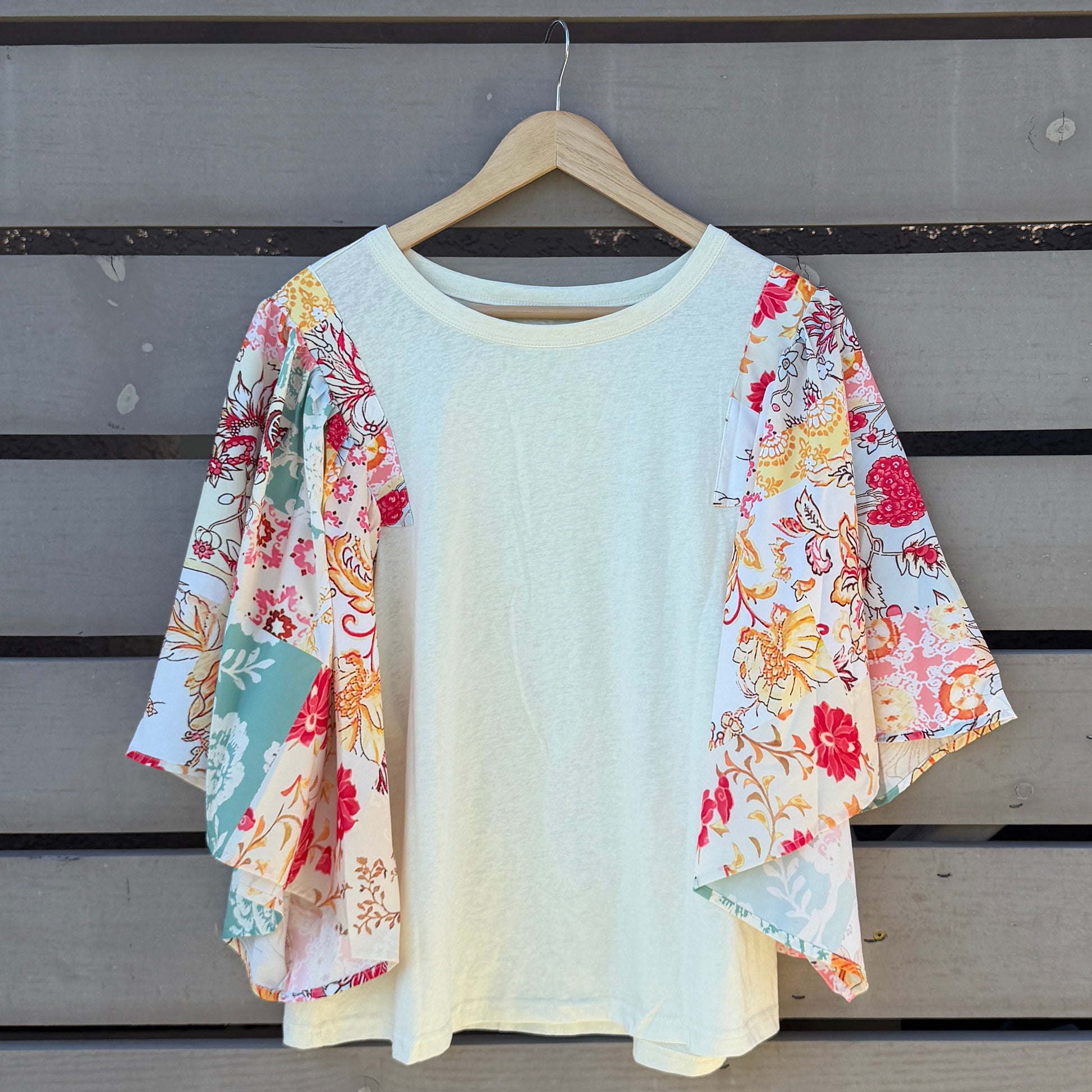 White blouse with floral sleeves on a wooden hanger against a wooden wall.