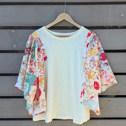 White blouse with floral sleeves on a wooden hanger against a wooden wall.