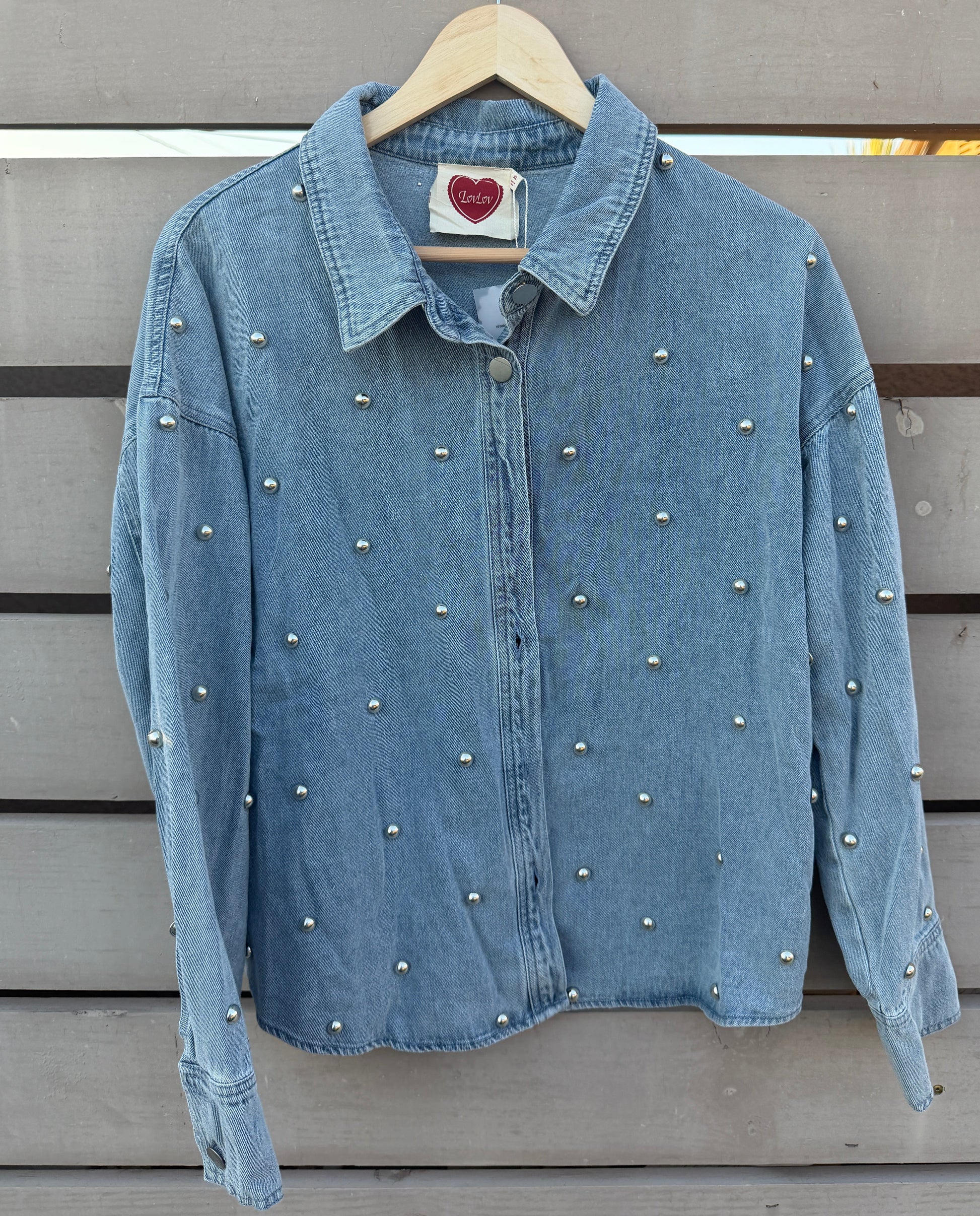 A light blue denim jacket decorated with small silver studs, hanging on a wooden hanger against a gray wooden slat wall.