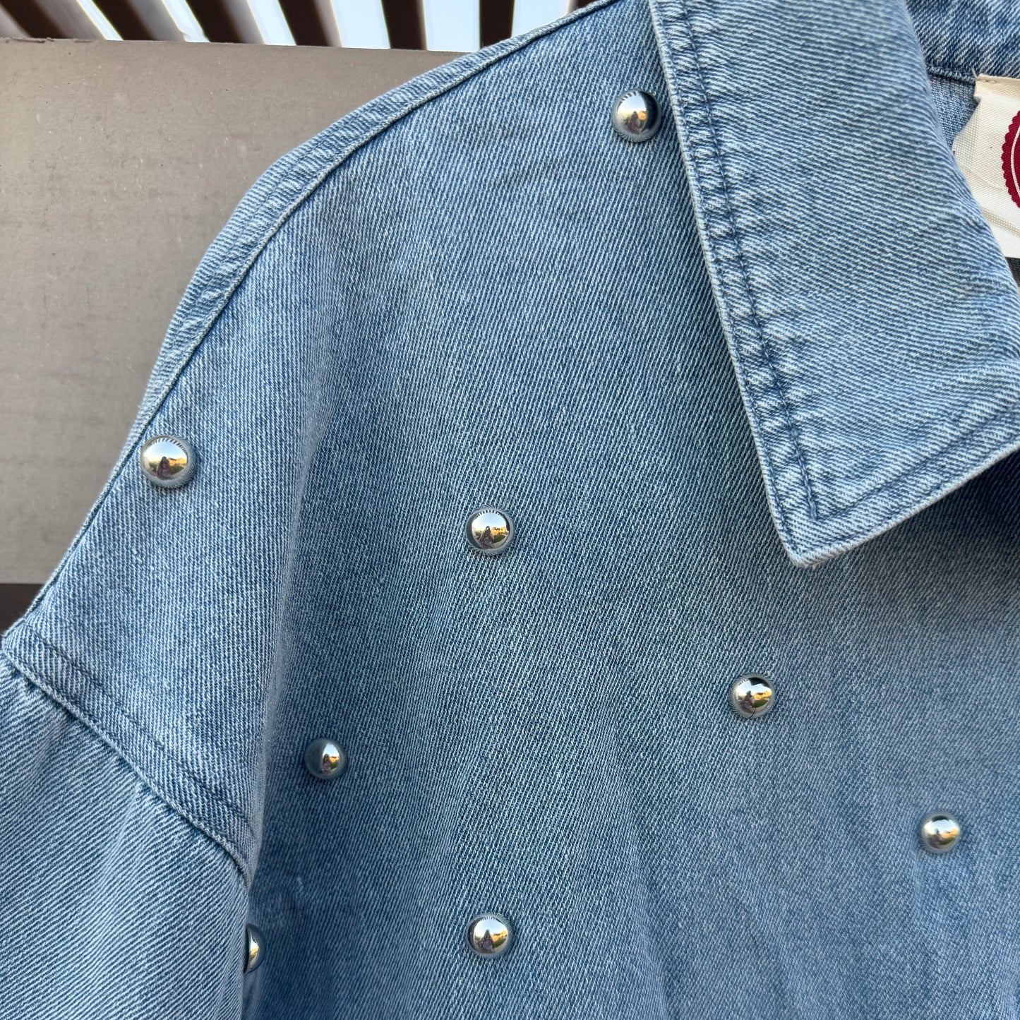A close-up of a light blue denim jacket decorated with small silver metallic studs, showing the collar and sleeve details.