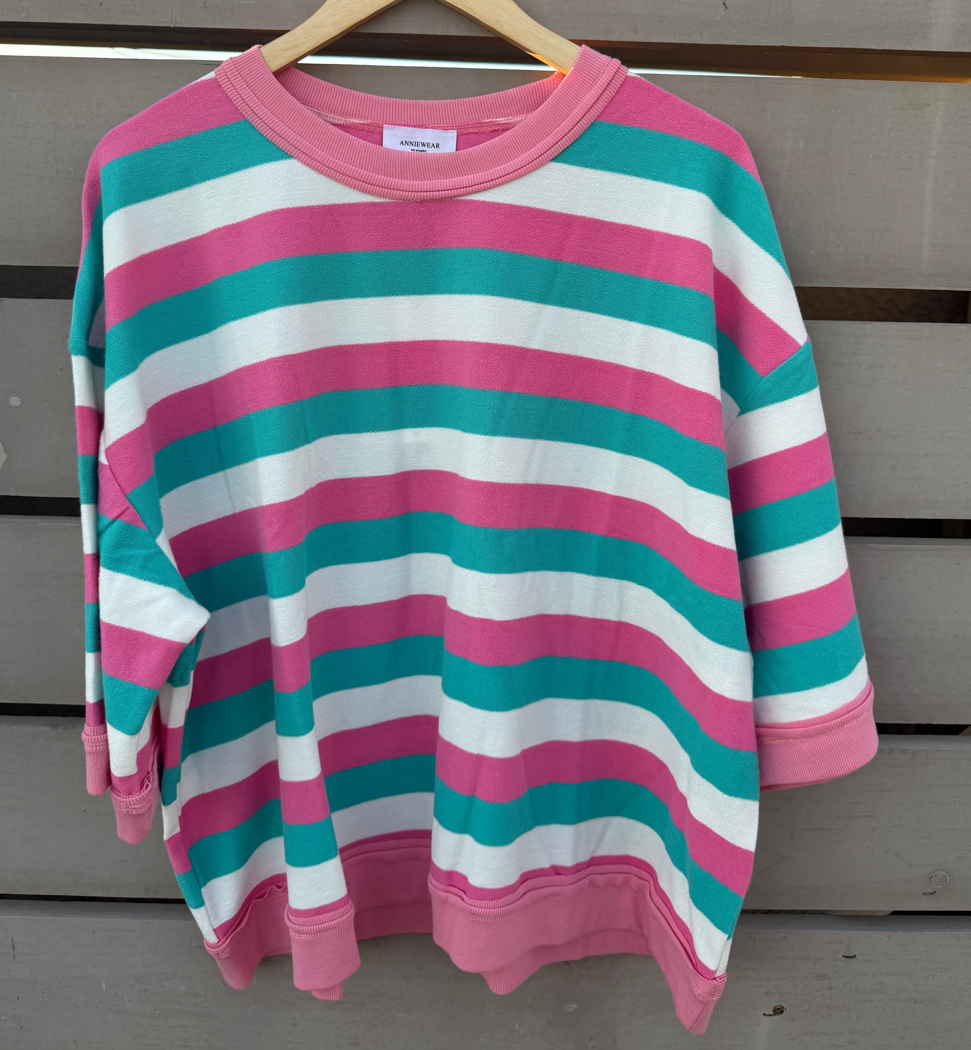 A pink, teal, and white striped long-sleeve crewneck sweatshirt hanging on a wooden hanger against a gray wooden slat wall.