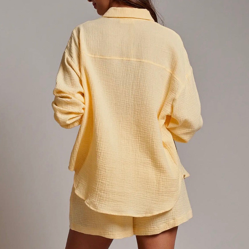 A woman wearing a pale yellow textured two-piece lounge set consisting of a long-sleeve button-up shirt and matching shorts, shown from a rear three-quarter view against a neutral gray studio background.