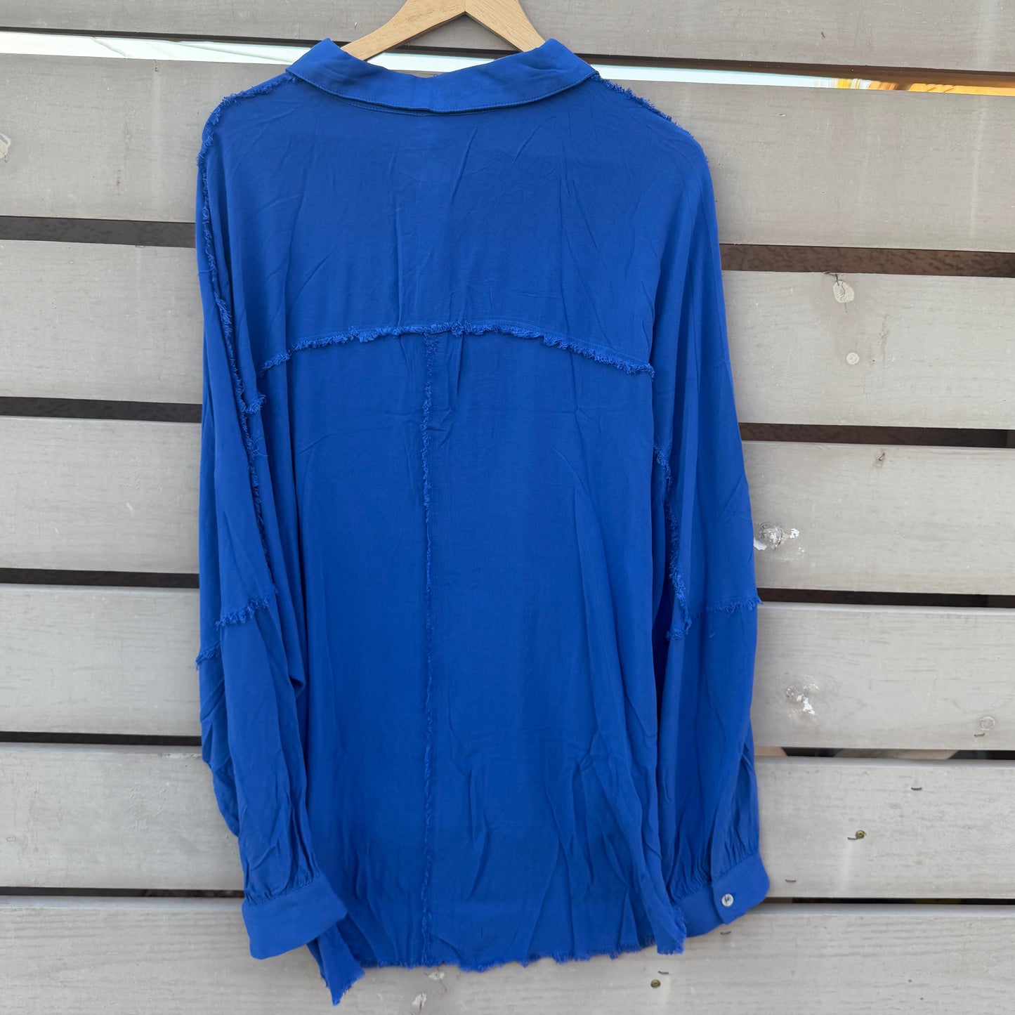 A royal blue long-sleeve blouse with a relaxed, slightly distressed fit, hanging on a wooden hanger against a light gray wooden slat wall.