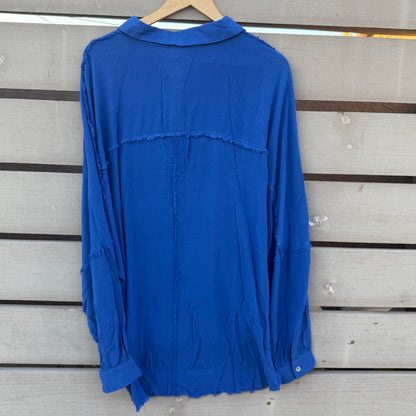 A royal blue long-sleeve blouse with a relaxed, slightly distressed fit, hanging on a wooden hanger against a light gray wooden slat wall.