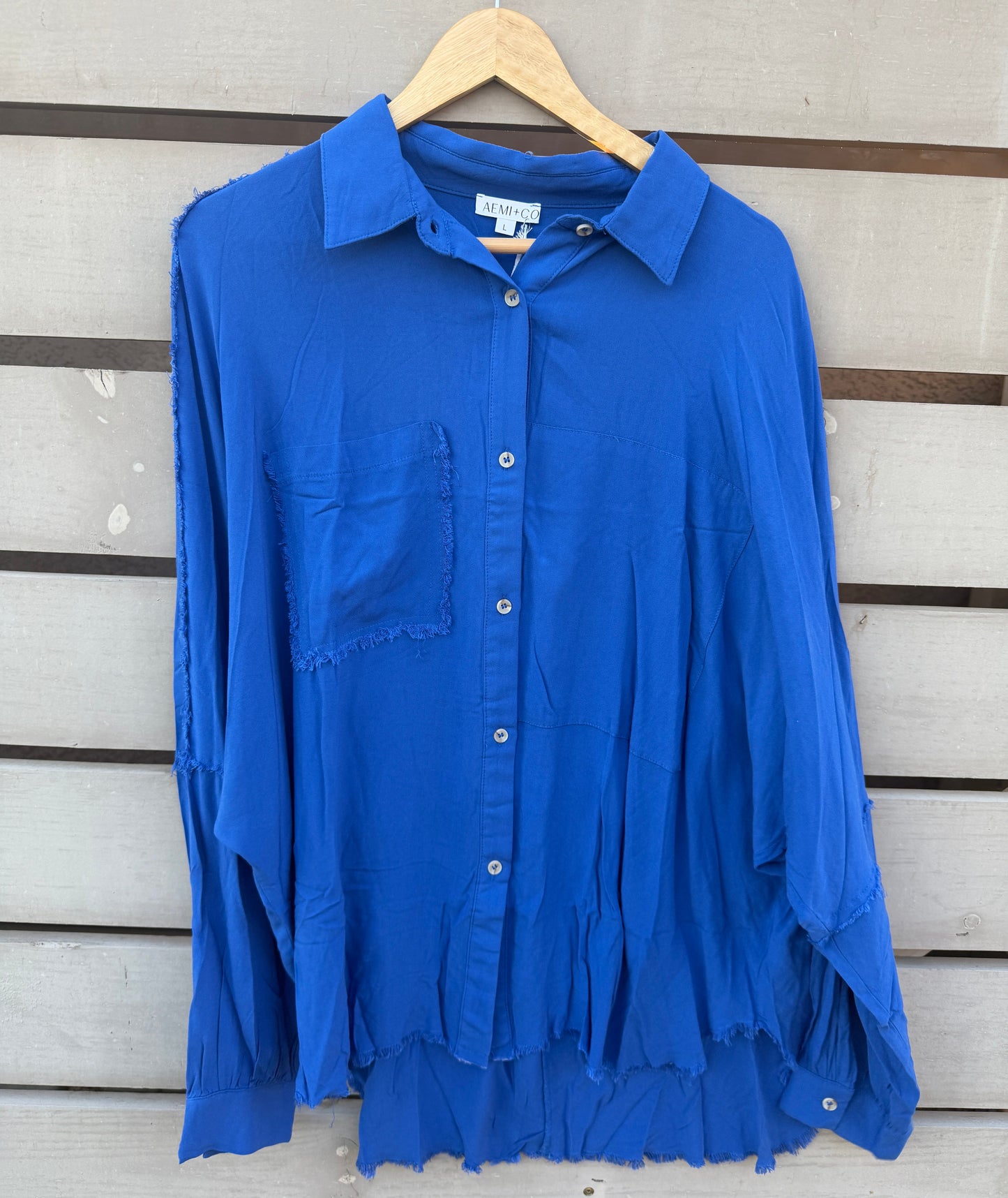 A bright royal blue long-sleeve button-up shirt with a chest pocket and raw, frayed harem-style hem, hanging on a wooden hanger against a light gray wooden slat wall.
