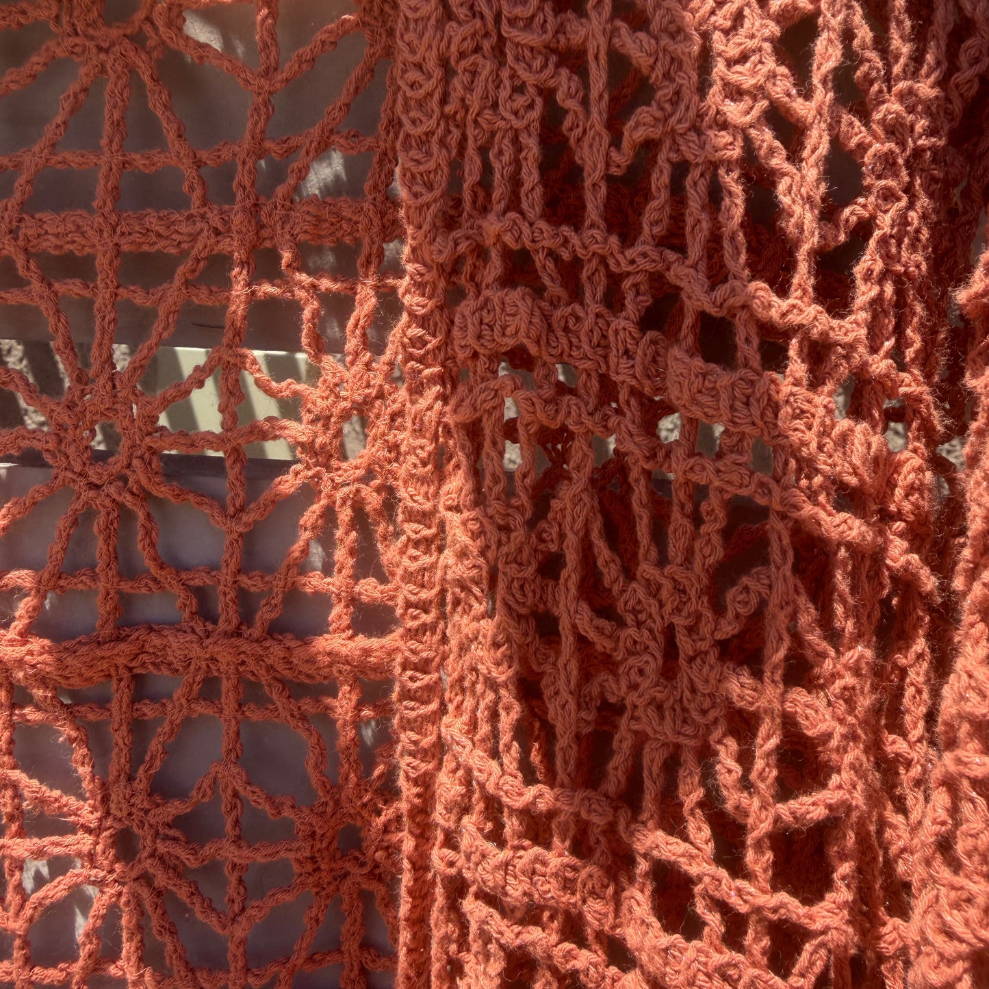 A close-up of a rust-orange crochet or lace textile draped over a wooden rod, showing the openwork pattern and texture in bright natural light.
