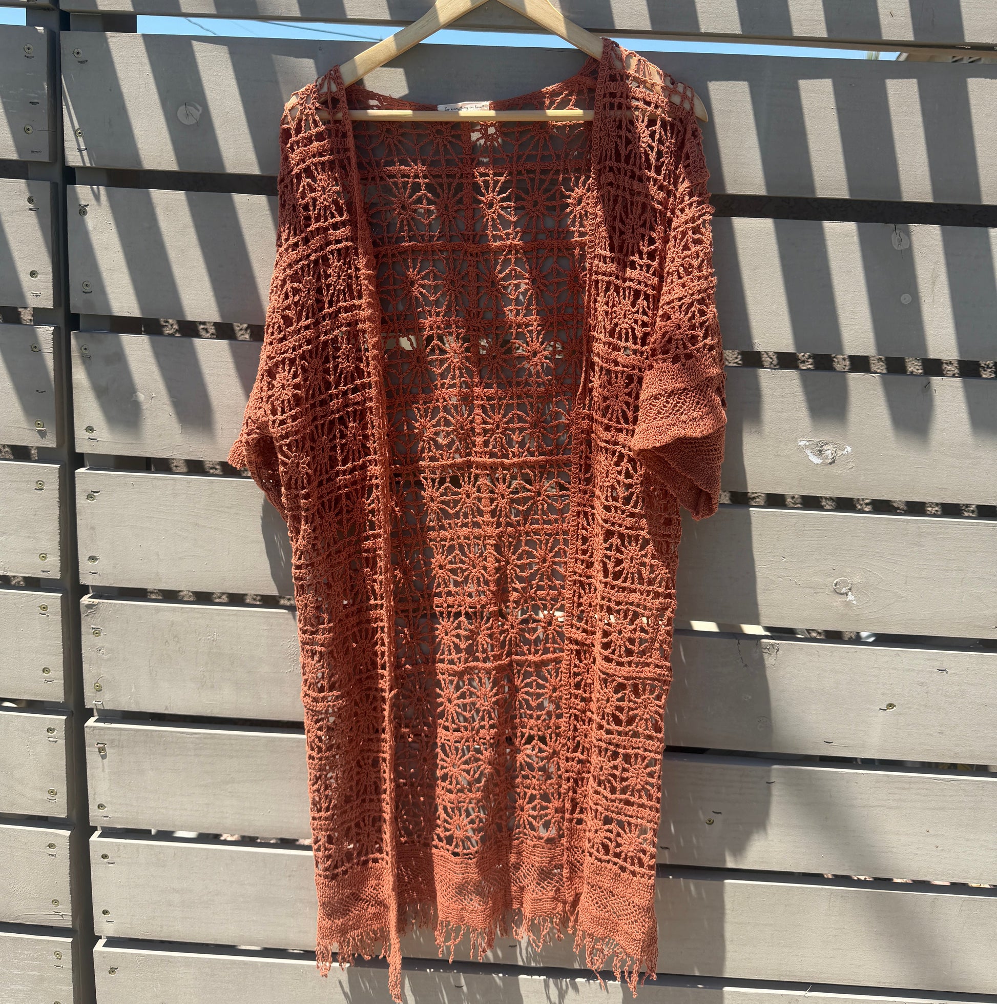 A terracotta orange crochet knit cardigan with a textured openwork pattern, hanging on a light-colored wooden hanger against a gray slatted deck background.