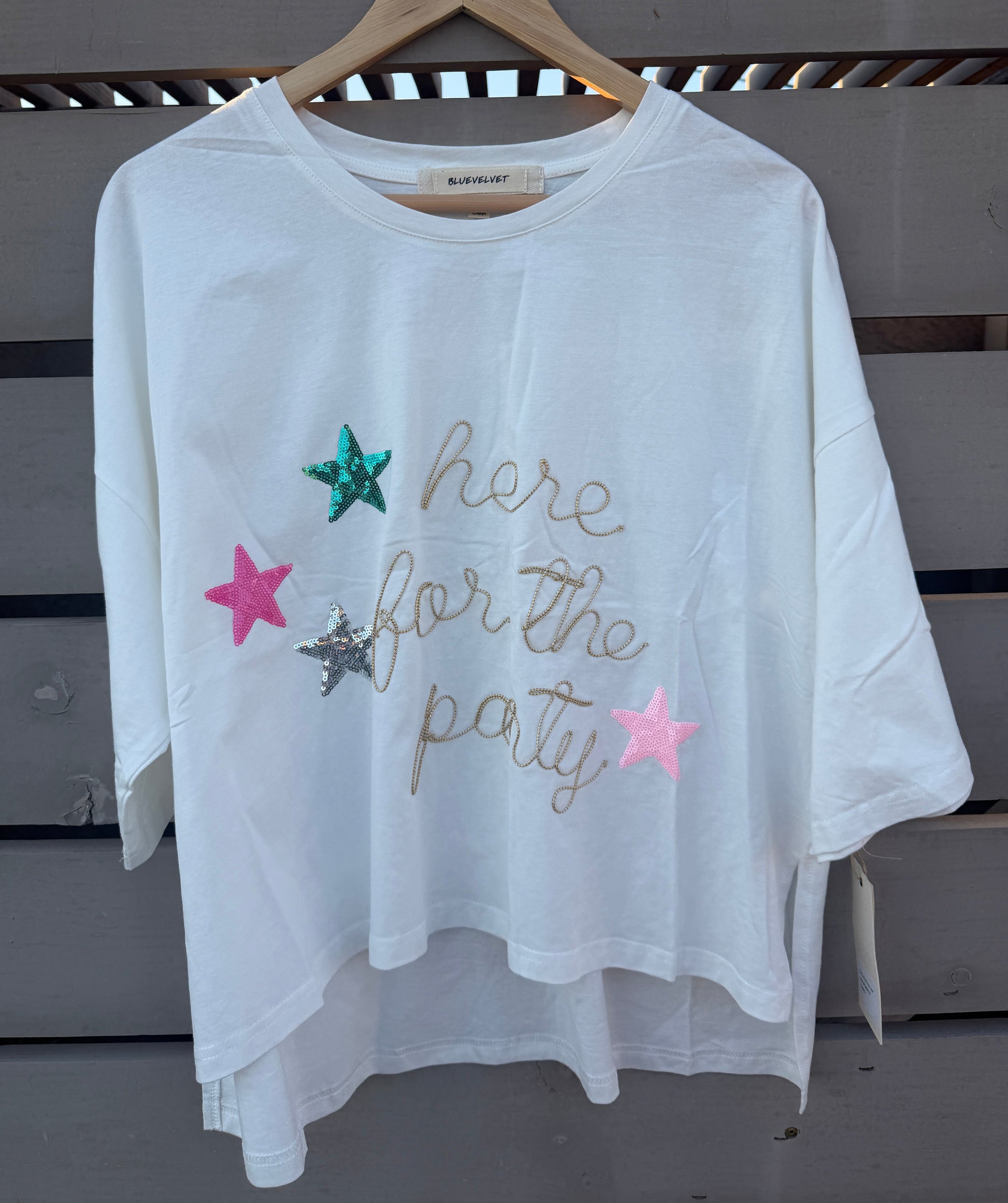 A white short-sleeve t-shirt with 'here for the party' written in gold glitter script and decorated with three glittery star accents (green, pink, and silver), hanging on a wooden hanger against a gray wooden slat wall.