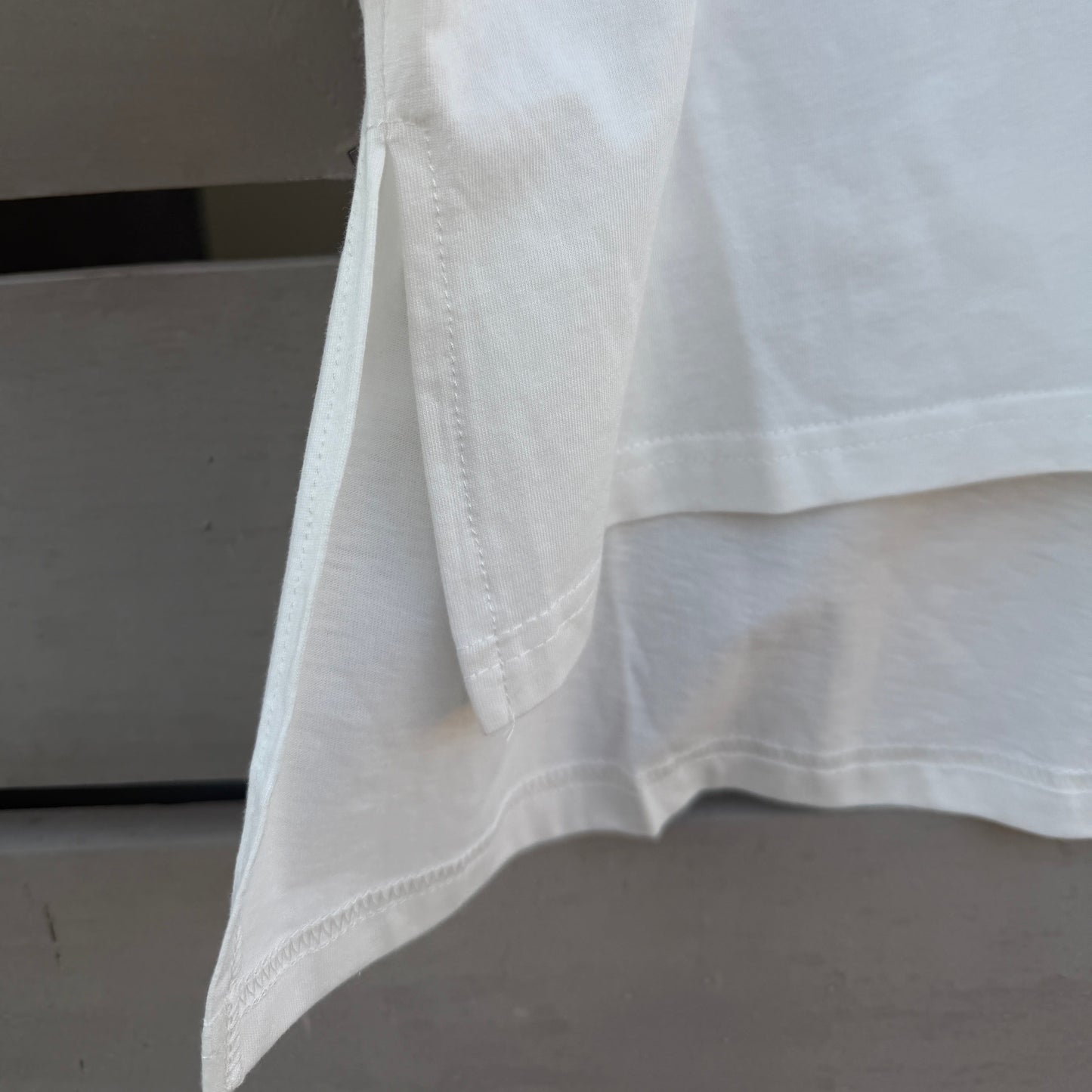 Close-up of a white high-low hem t-shirt, showing the folded hemline and clean stitching, with a small colorful graphic detail visible in the upper right corner.
