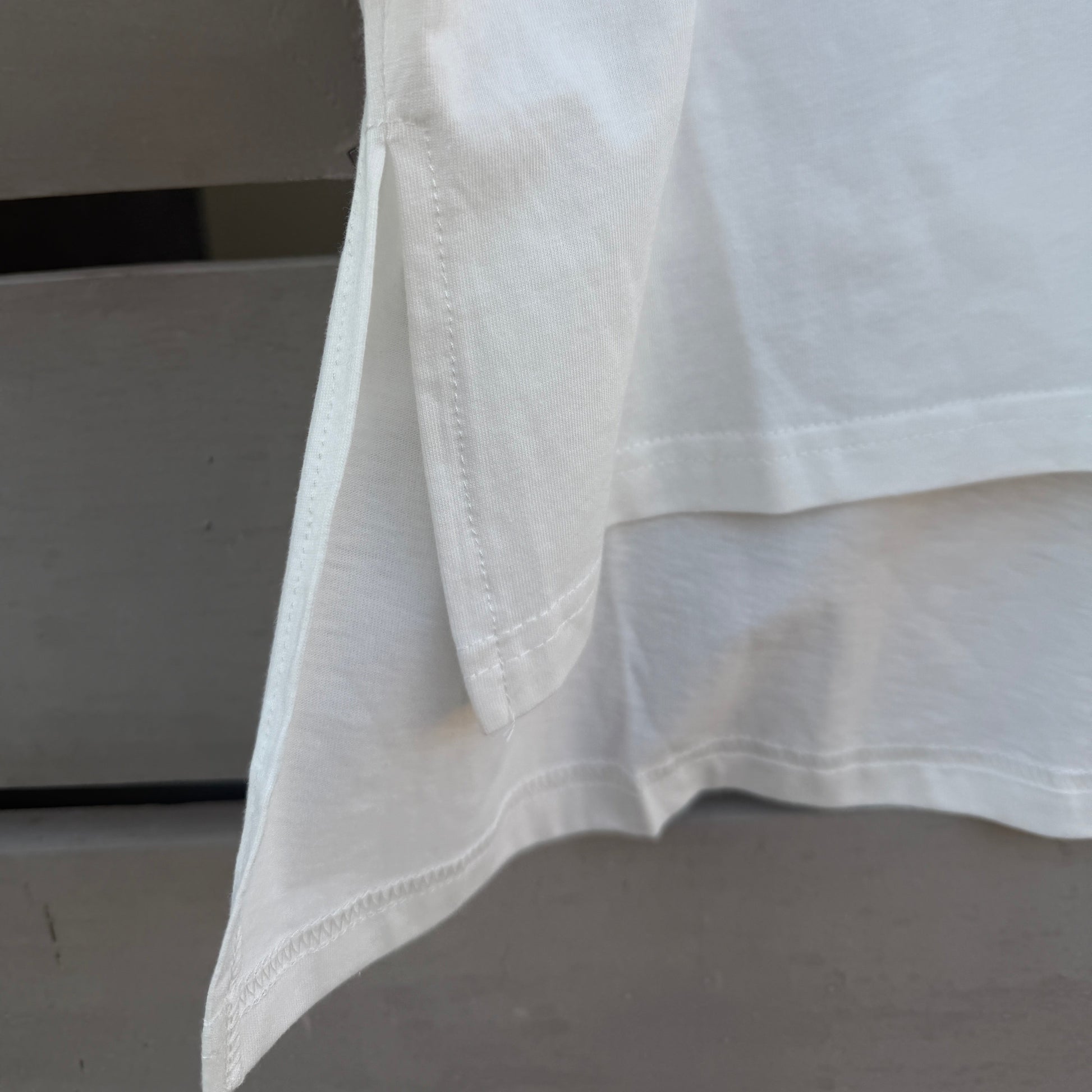 Close-up of a white high-low hem t-shirt, showing the folded hemline and clean stitching, with a small colorful graphic detail visible in the upper right corner.