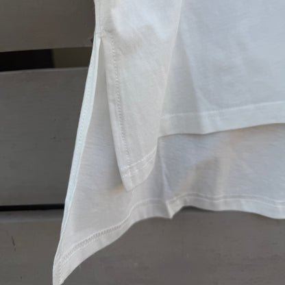 Close-up of a white high-low hem t-shirt, showing the folded hemline and clean stitching, with a small colorful graphic detail visible in the upper right corner.
