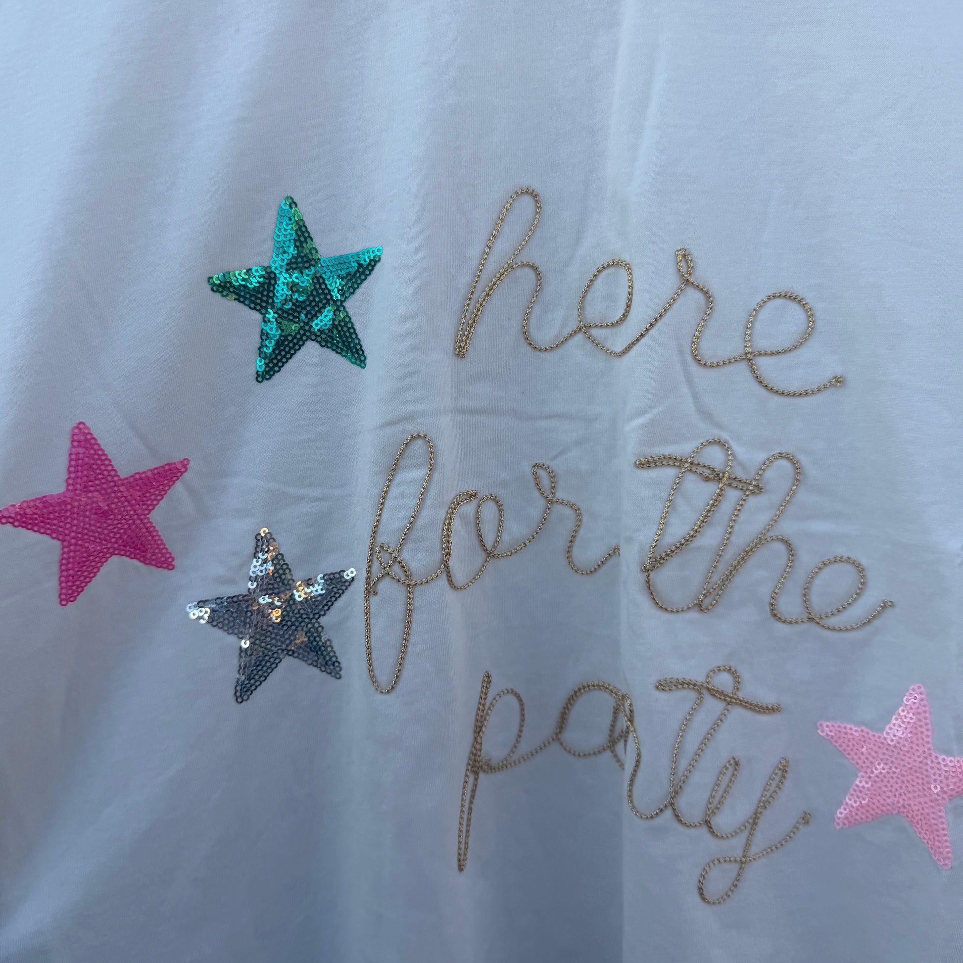 A white t-shirt featuring the phrase 'here for the party' in gold glitter script, decorated with three sequined stars in teal, pink, and silver.