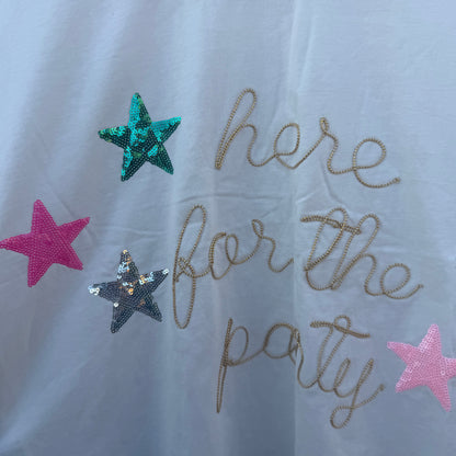 A white t-shirt featuring the phrase 'here for the party' in gold glitter script, decorated with three sequined stars in teal, pink, and silver.