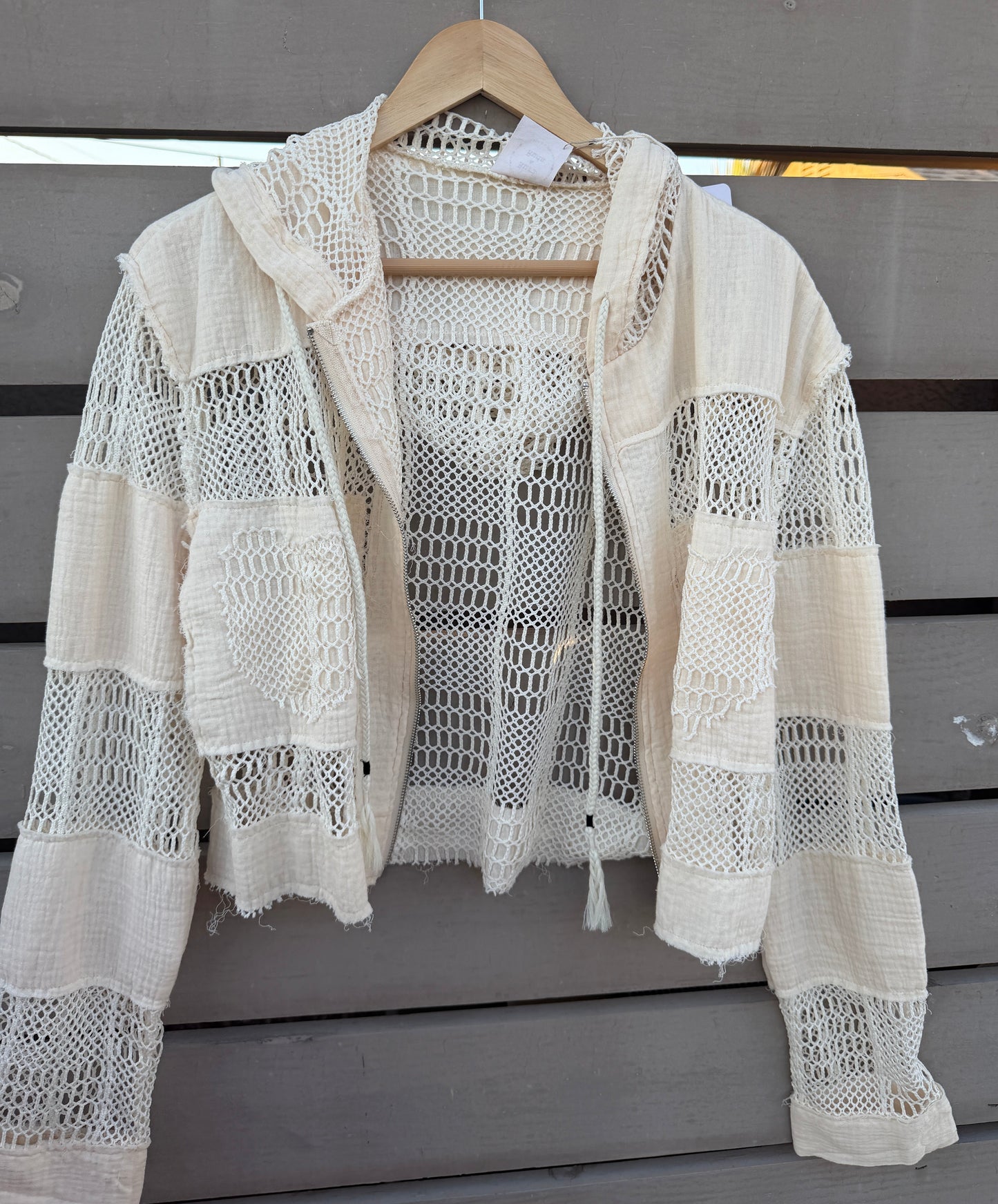 An off-white crochet knit hooded cardigan with distressed details, hanging on a wooden hanger against a gray wooden slat wall.
