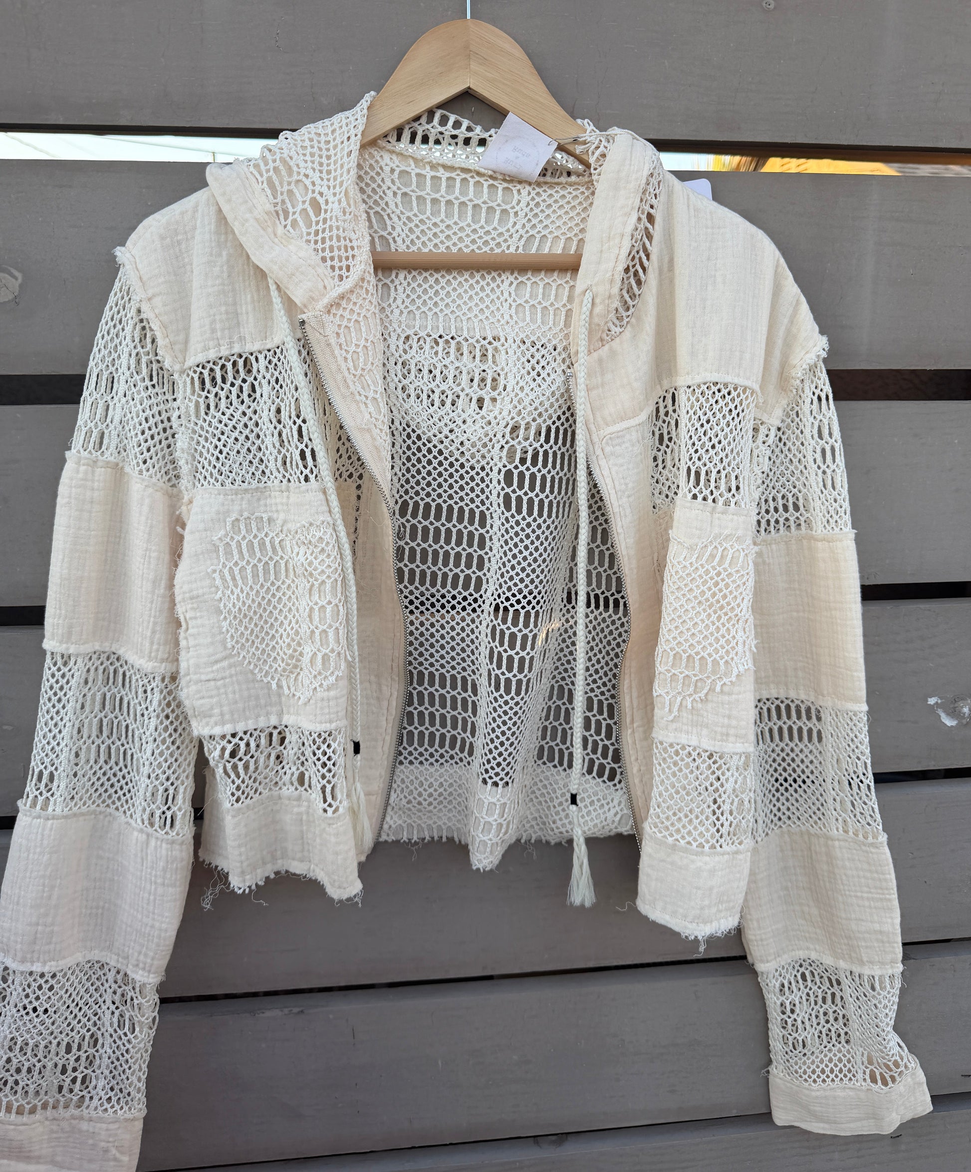 An off-white crochet knit hooded cardigan with distressed details, hanging on a wooden hanger against a gray wooden slat wall.