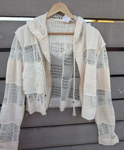 An off-white crochet knit hooded cardigan with distressed details, hanging on a wooden hanger against a gray wooden slat wall.