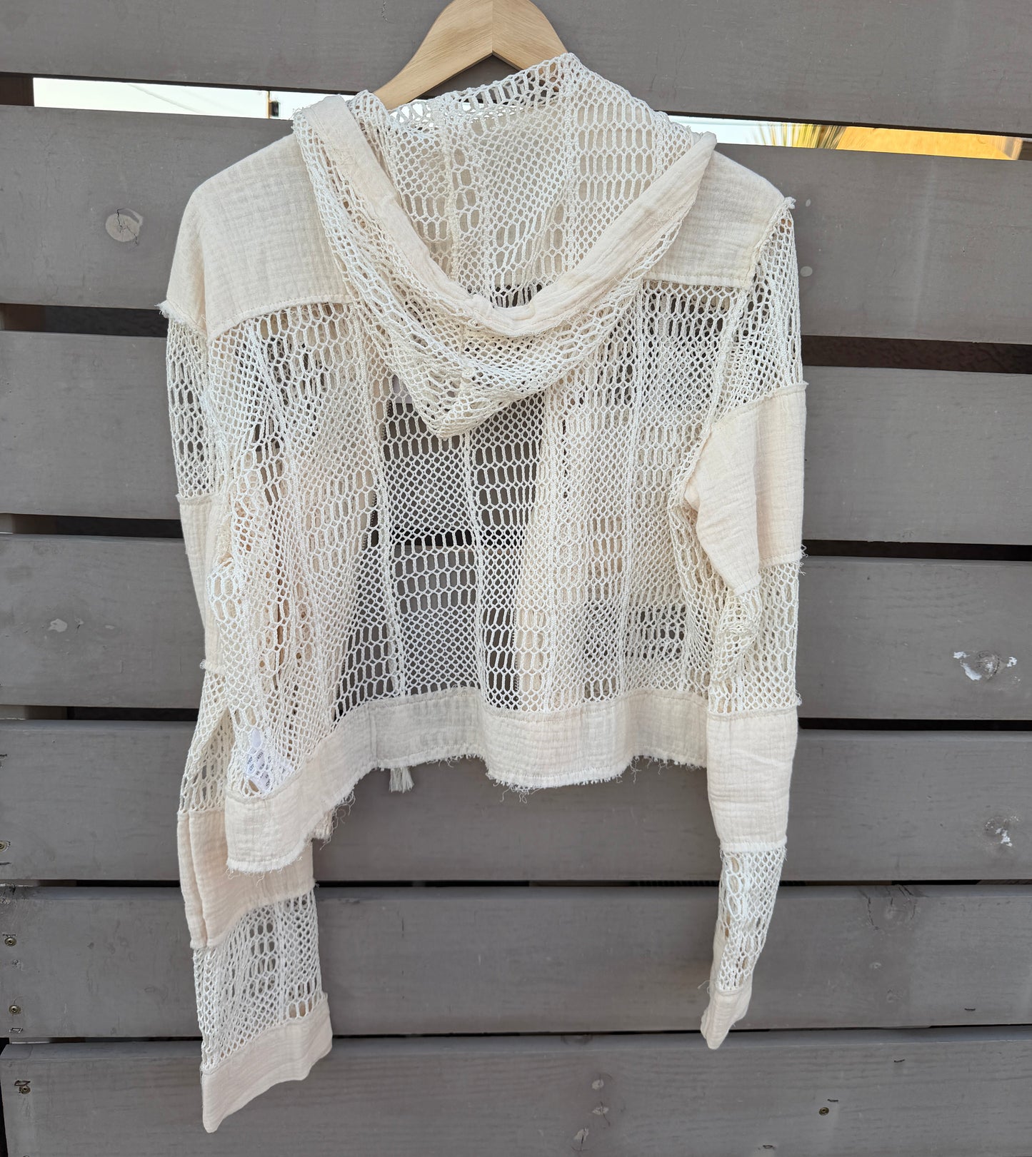 An off-white crochet knit hooded cardigan with long sleeves, hanging on a wooden hanger against a gray wooden slat wall.