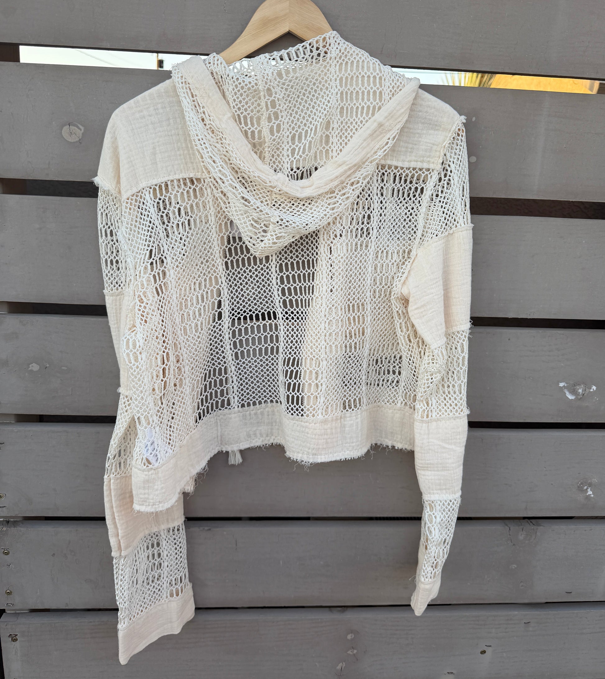An off-white crochet knit hooded cardigan with long sleeves, hanging on a wooden hanger against a gray wooden slat wall.