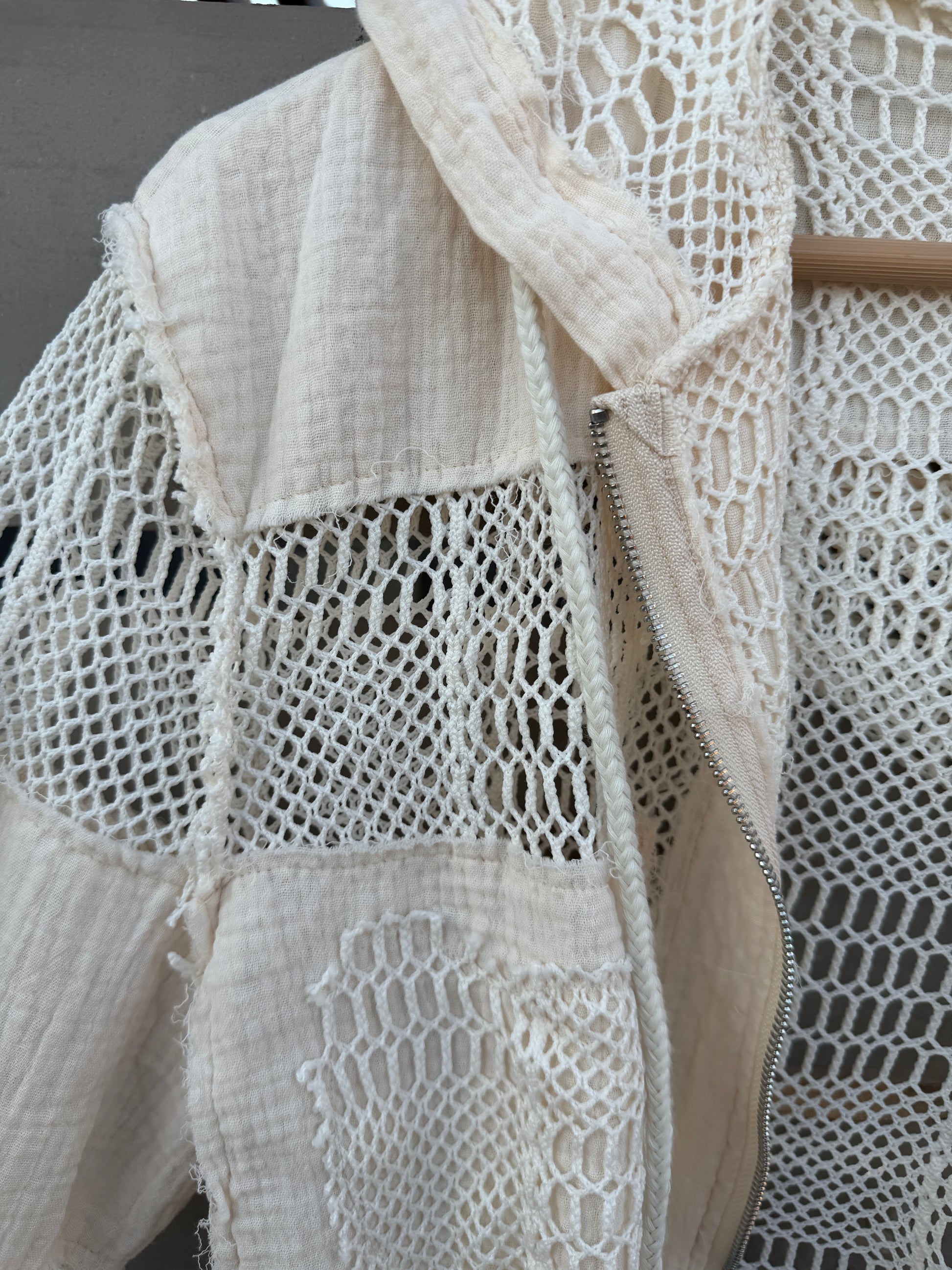 Close-up of a cream-colored patchwork jacket featuring textured woven panels and a central zipper, shown hanging on a wooden hanger.