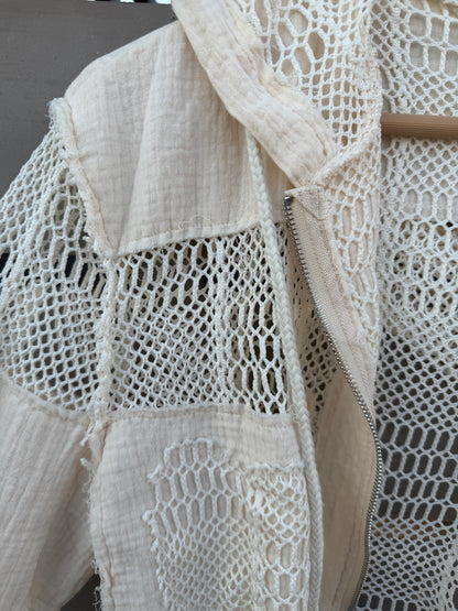 Close-up of a cream-colored patchwork jacket featuring textured woven panels and a central zipper, shown hanging on a wooden hanger.