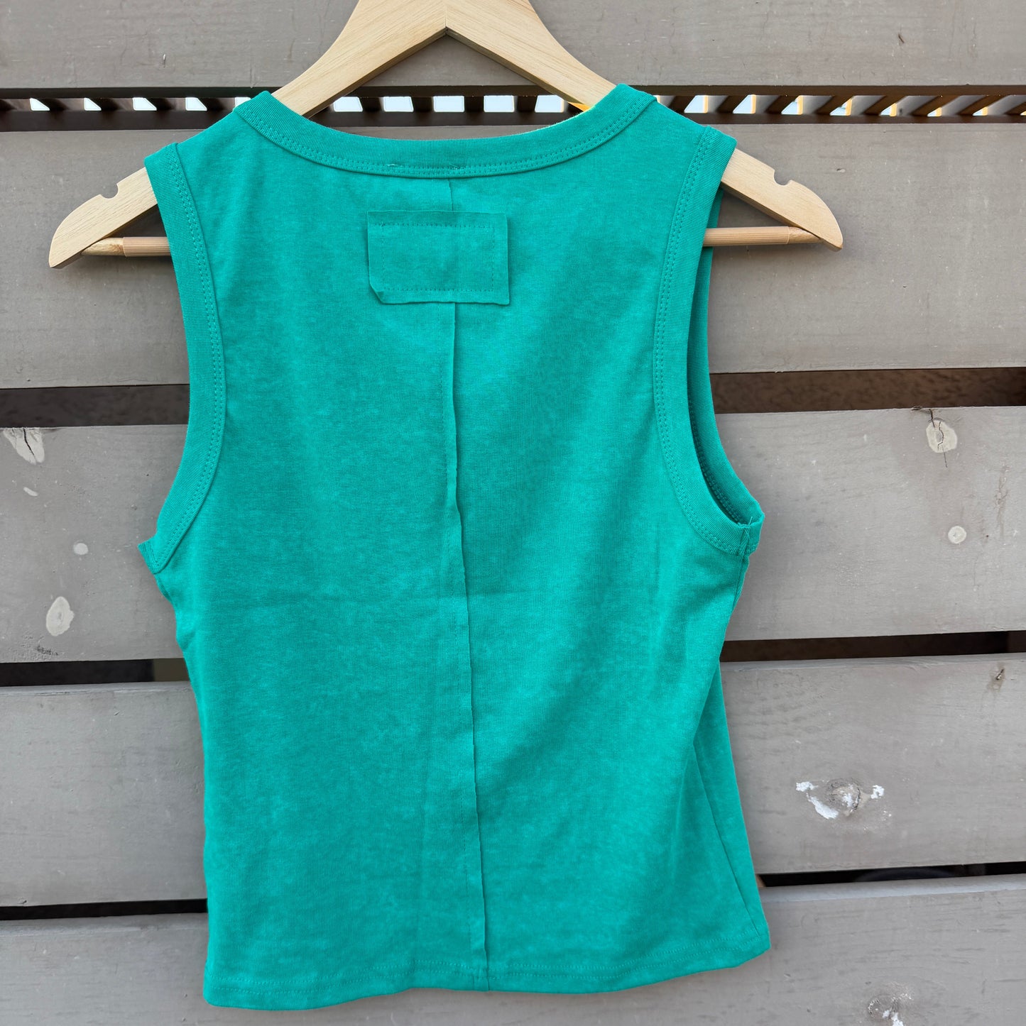 A sleeveless teal green top hanging on a wooden hanger against a gray wooden slat wall.