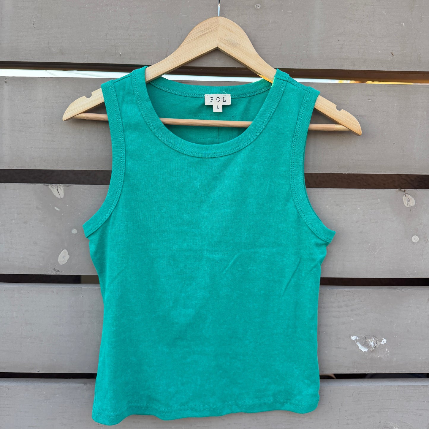 A bright teal green sleeveless tank top hanging on a wooden hanger against a gray wooden slat wall.