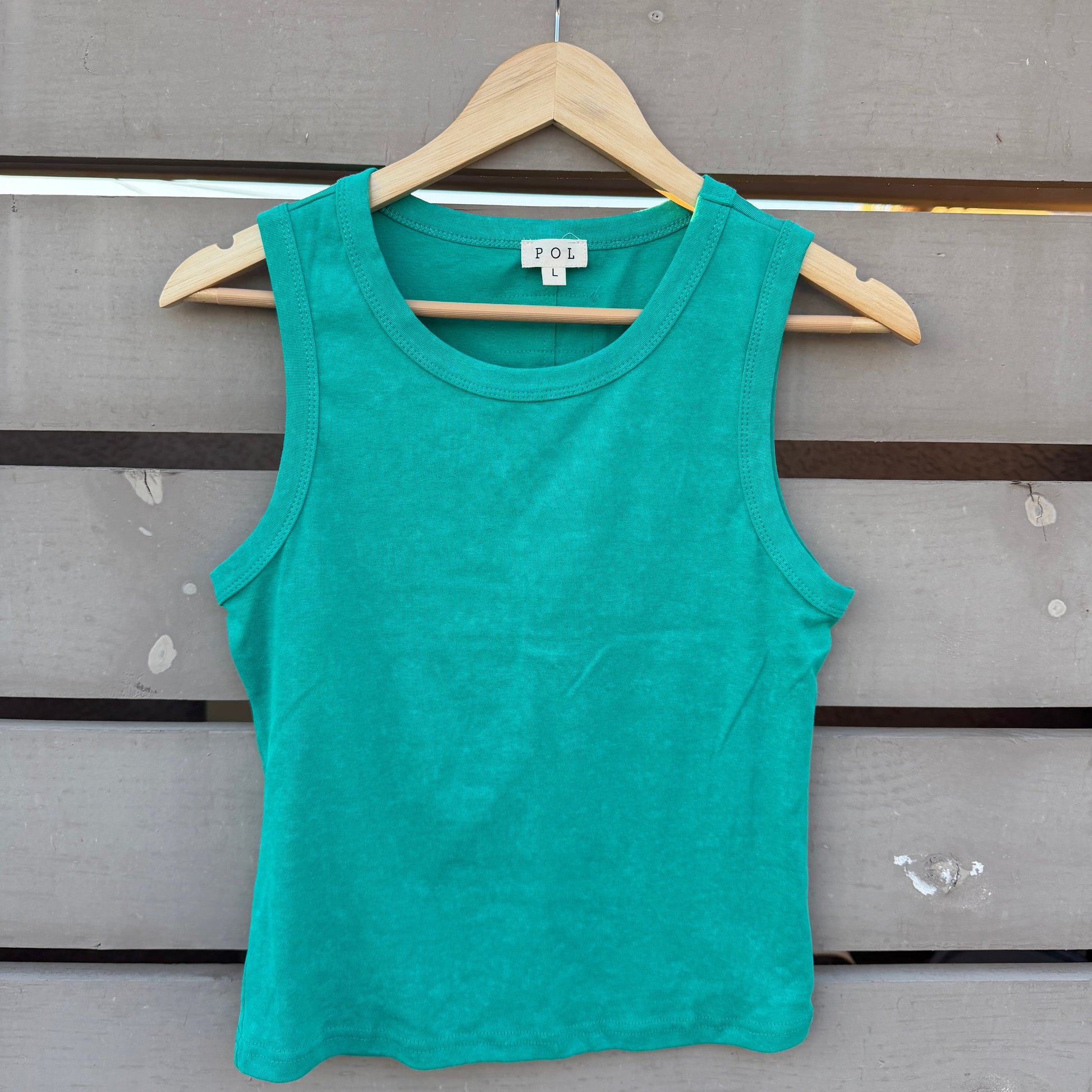 A bright teal green sleeveless tank top hanging on a wooden hanger against a gray wooden slat wall.