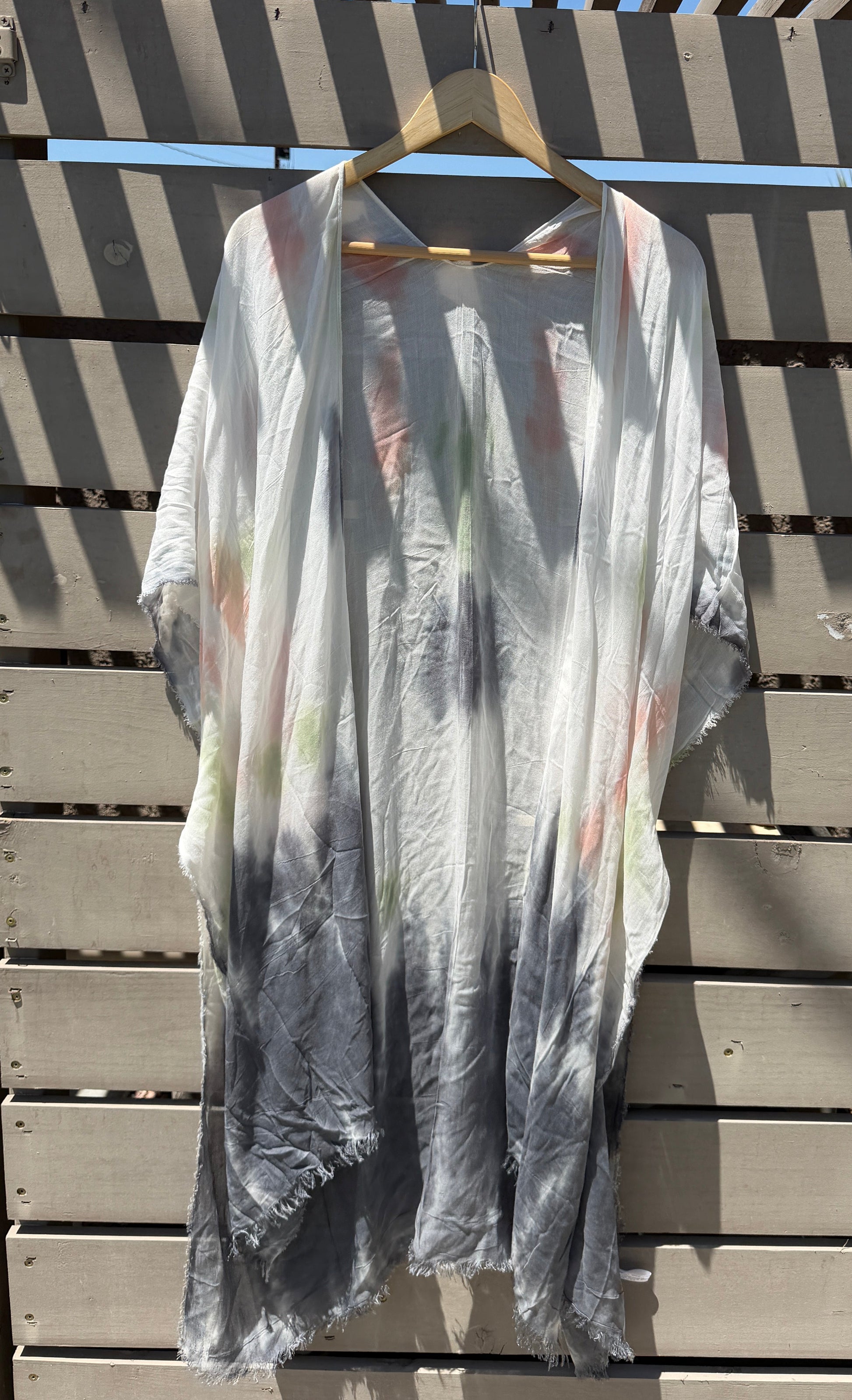 A lightweight, semi-sheer kaftan-style cover-up dress with a gray and white tie-dye pattern, hanging on a wooden hanger against a wooden slat wall under a slatted roof.