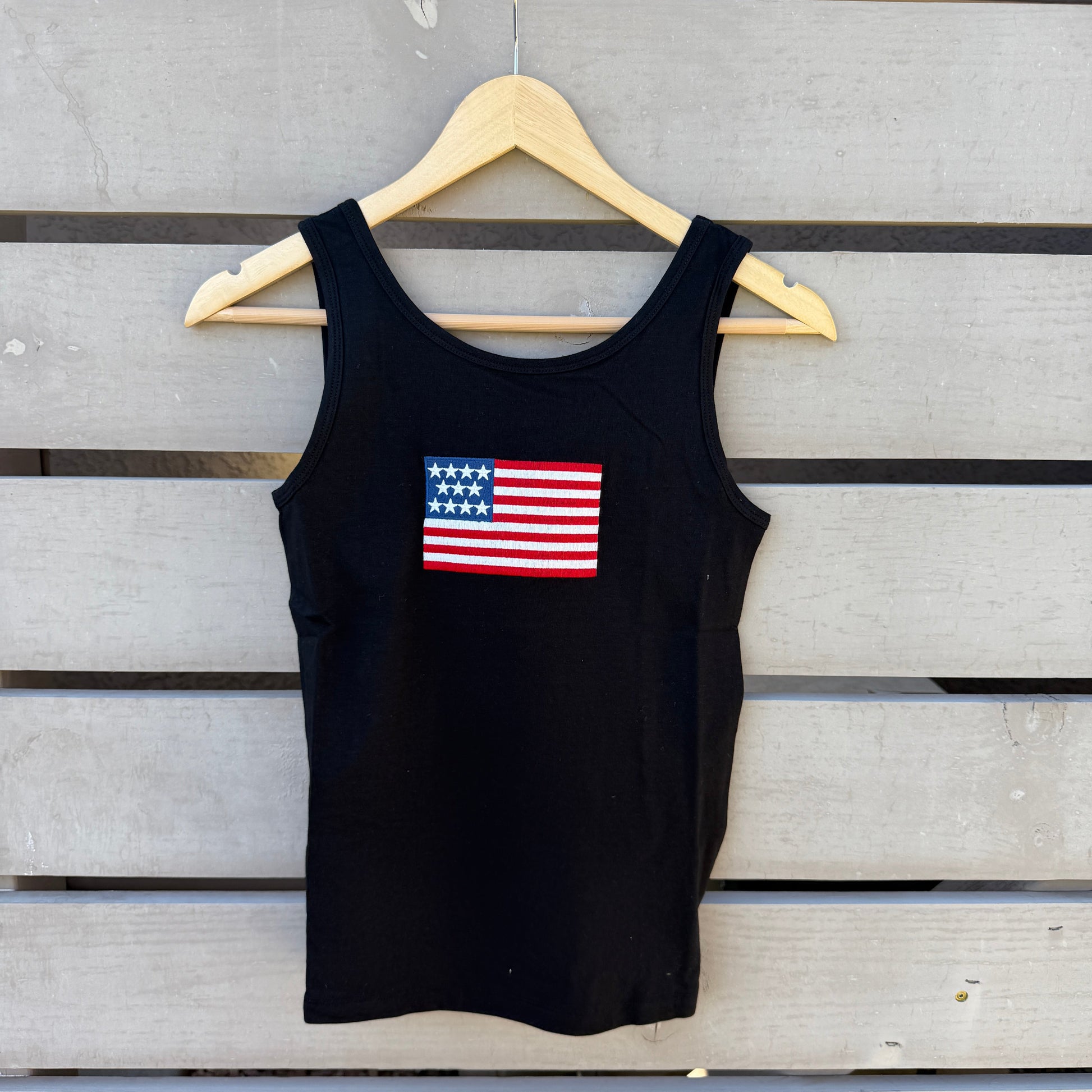 Black tank top with American flag design on a wooden hanger against a wooden slat background