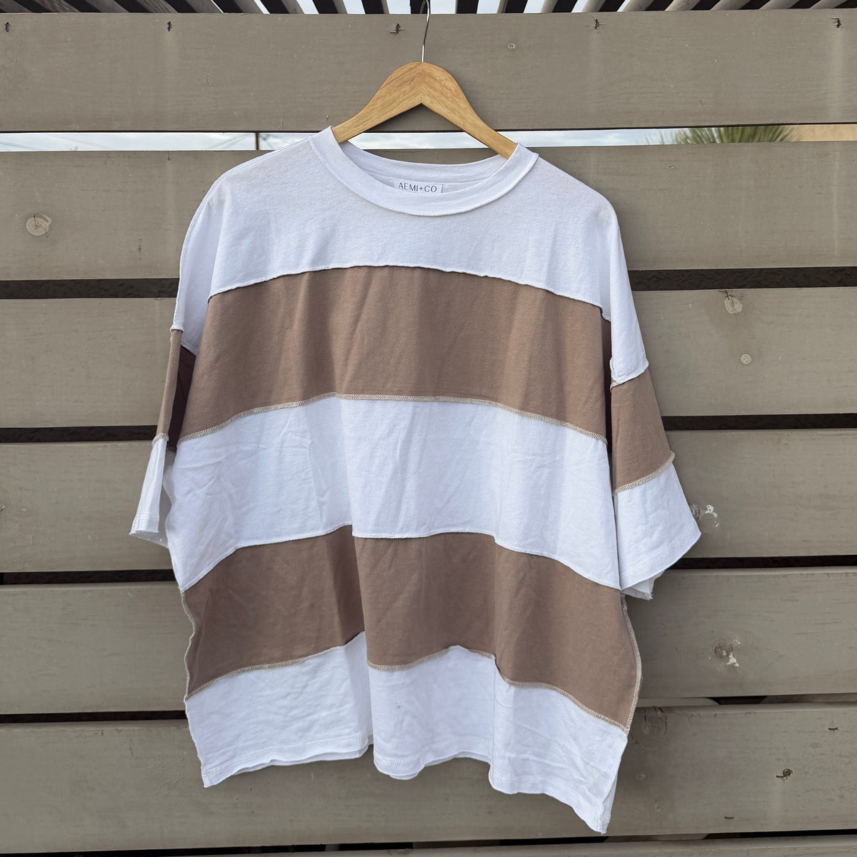 A loose short-sleeve striped T-shirt hanging on a wooden hanger against a gray wooden slat fence outdoors.