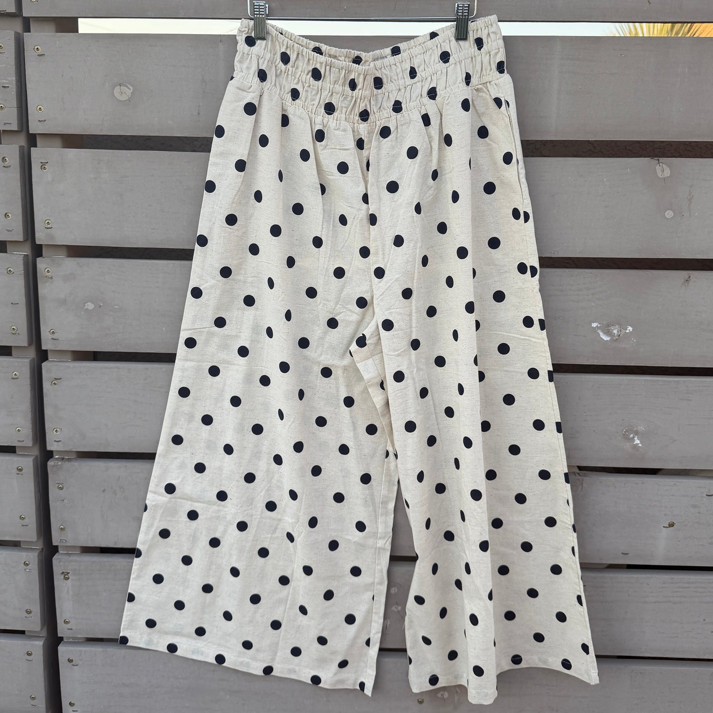 White wide-leg cropped pants with black polka dots, hanging on a wooden hanger against a gray wooden fence.
