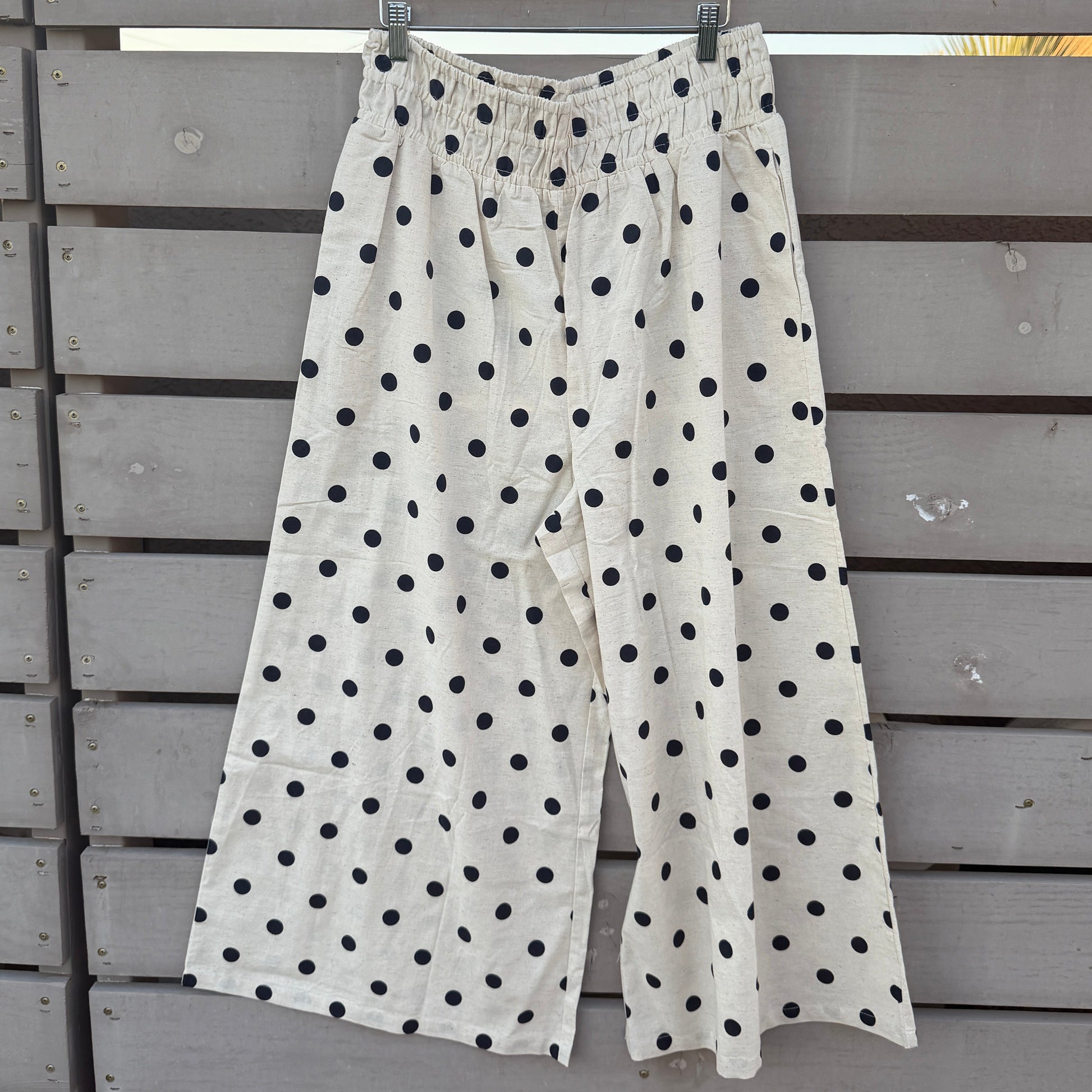 White wide-leg cropped pants with black polka dots, hanging on a wooden hanger against a gray wooden fence.