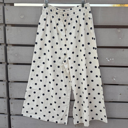 White wide-leg cropped pants with black polka dots, hanging on a wooden hanger against a gray wooden fence.