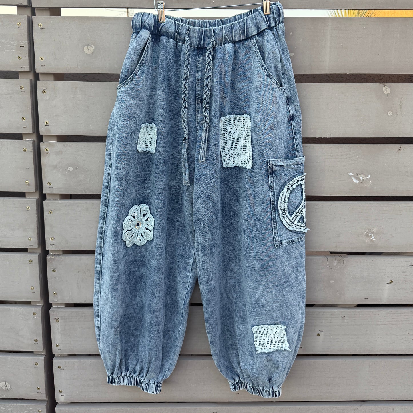 A pair of light blue washed denim harem-style pants with decorative patchwork details, hanging on a wooden hanger against a gray wooden slat fence.
