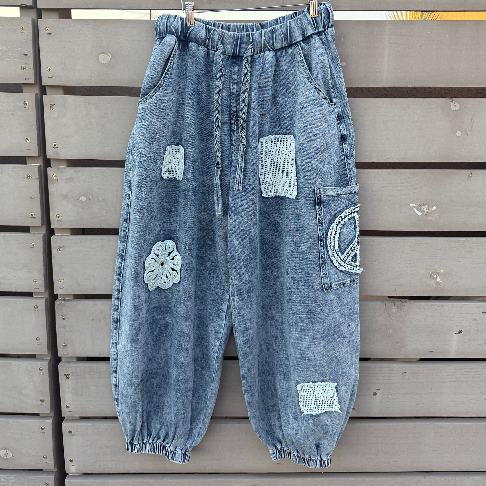 A pair of light blue washed denim harem-style pants with decorative patchwork details, hanging on a wooden hanger against a gray wooden slat fence.