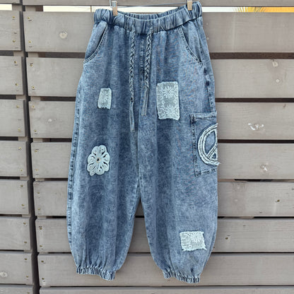 A pair of light blue washed denim harem-style pants with decorative patchwork details, hanging on a wooden hanger against a gray wooden slat fence.