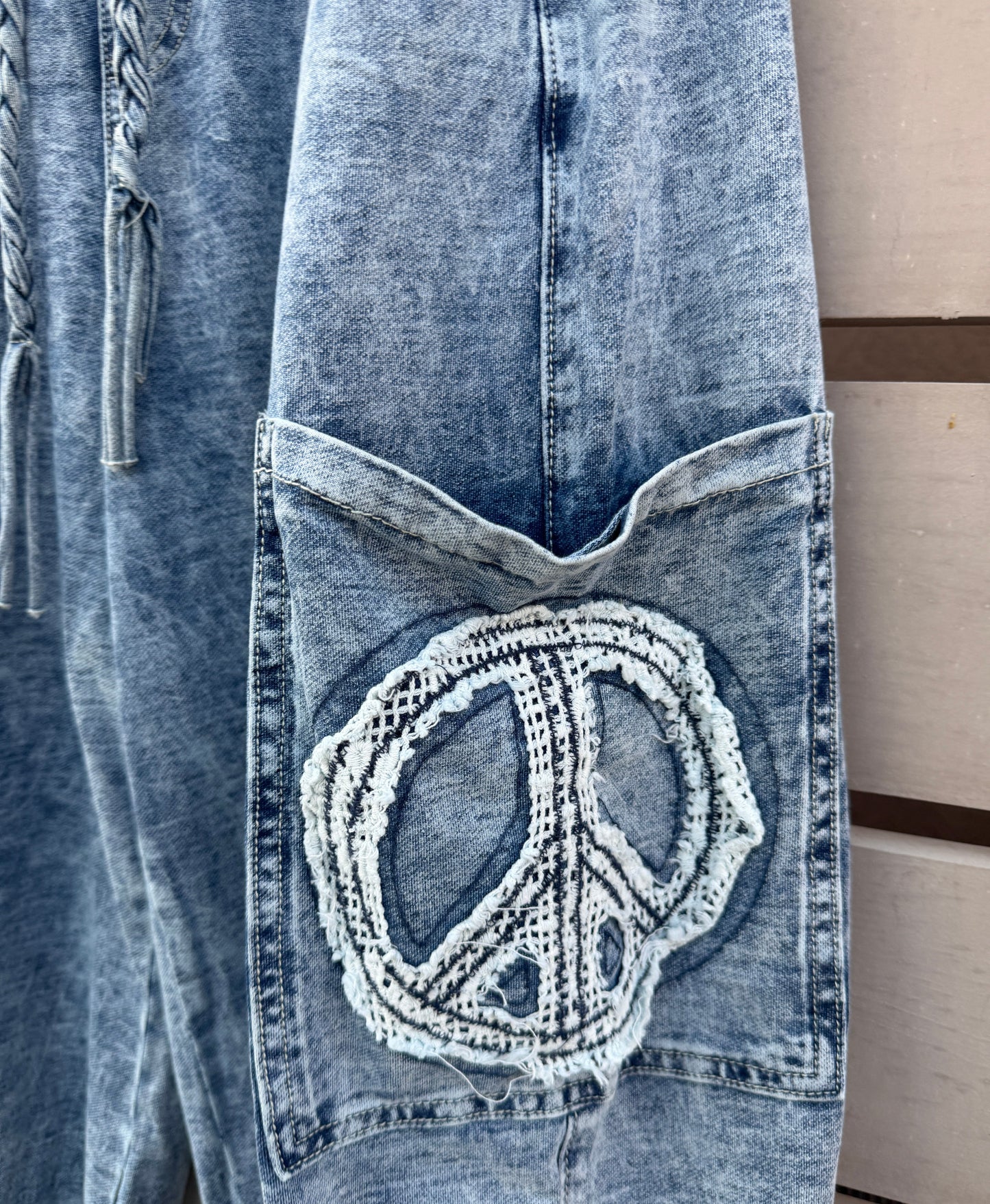 Close-up of a light blue denim garment featuring a large decorative patch on the pocket with a peace sign motif made from white crochet or knit fabric.