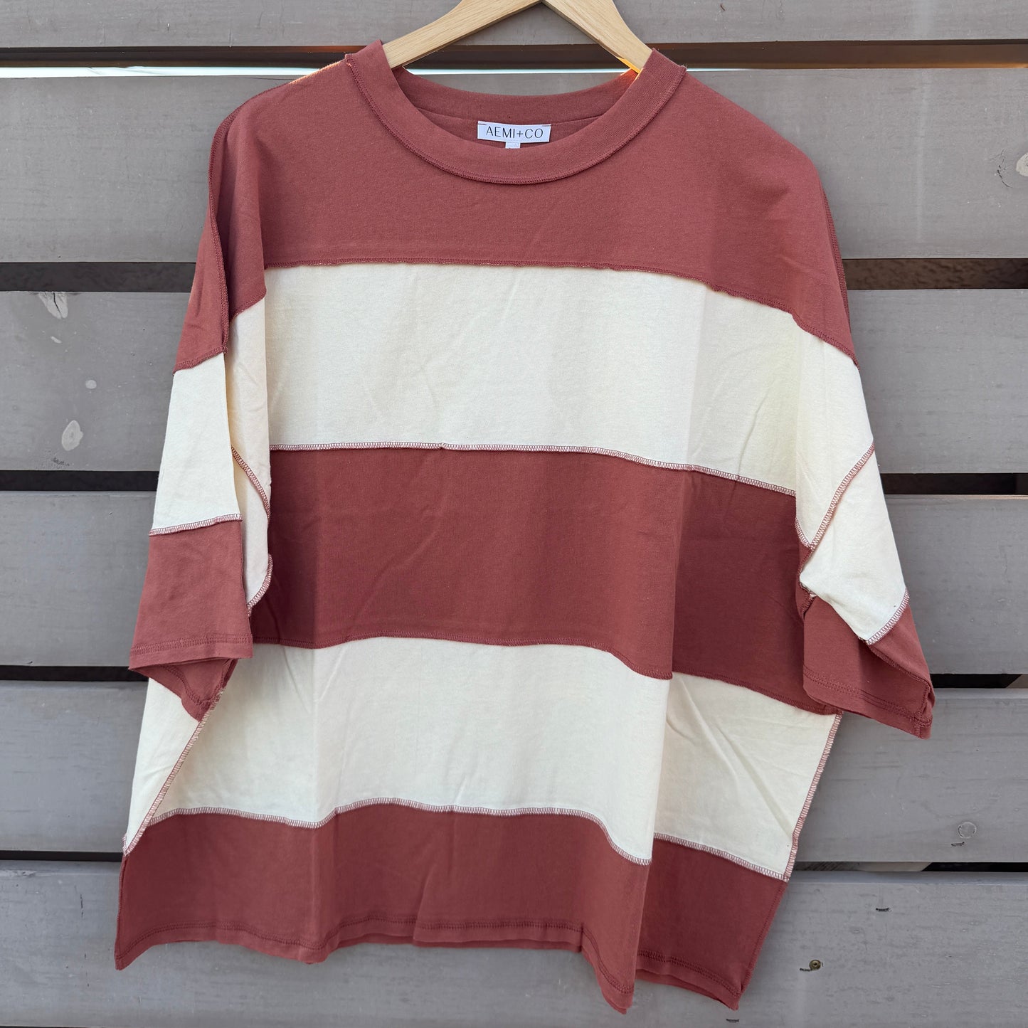 A short-sleeve striped t-shirt with horizontal bands of rust red and cream, hanging on a wooden hanger against a gray wooden slat wall.