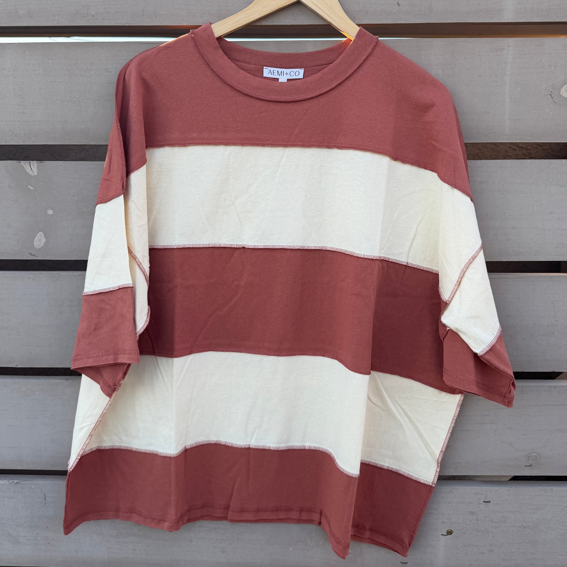 A short-sleeve striped t-shirt with horizontal bands of rust red and cream, hanging on a wooden hanger against a gray wooden slat wall.