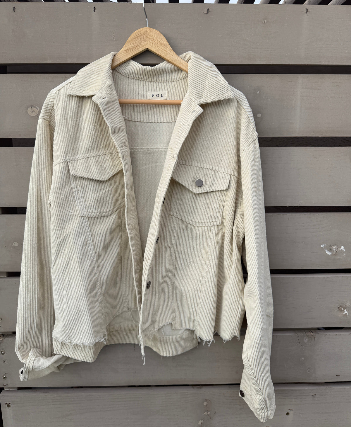 A light beige corduroy utility jacket with raw, distressed edges, hanging on a wooden hanger against a gray wooden slat wall. The jacket features a collar, button front, and flap chest pockets.