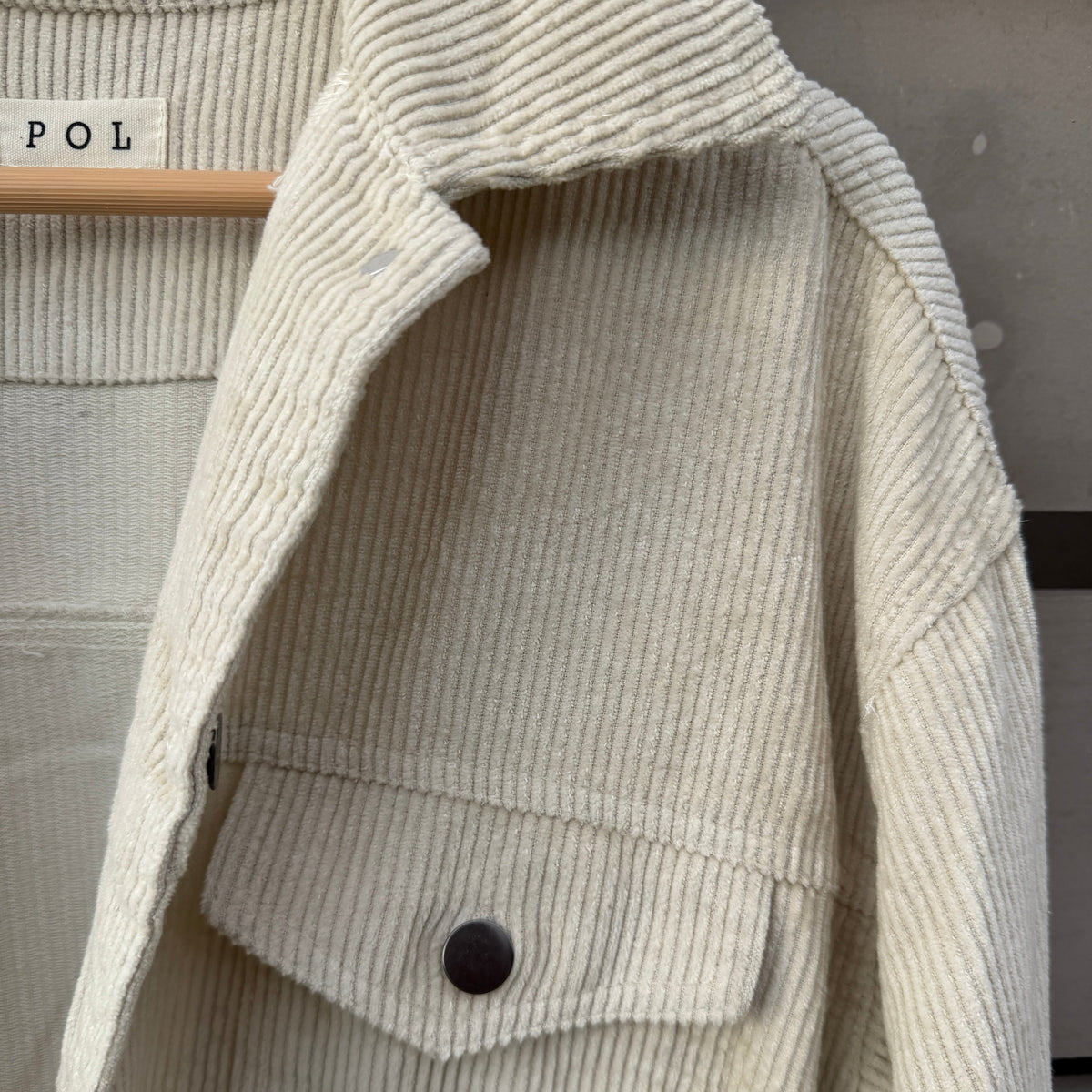 Close-up of a cream-colored ribbed corduroy jacket with a buttoned chest pocket, laid flat on a hanger.