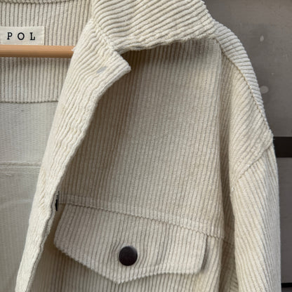 Close-up of a cream-colored ribbed corduroy jacket with a buttoned chest pocket, laid flat on a hanger.