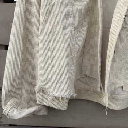 A close-up of a light beige ribbed corduroy jacket with raw, frayed hems, hanging against a weathered gray wooden background.