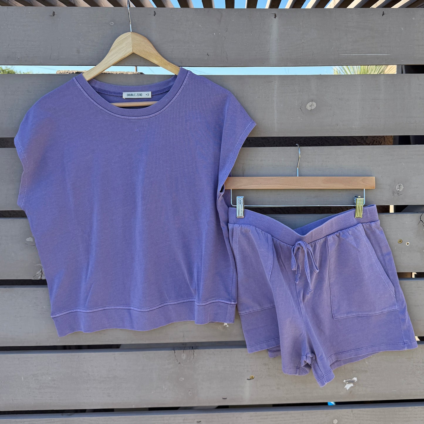 A lavender purple two-piece lounge set consisting of a short-sleeve cropped top and matching shorts, hanging on wooden hangers against a gray wooden slat wall.