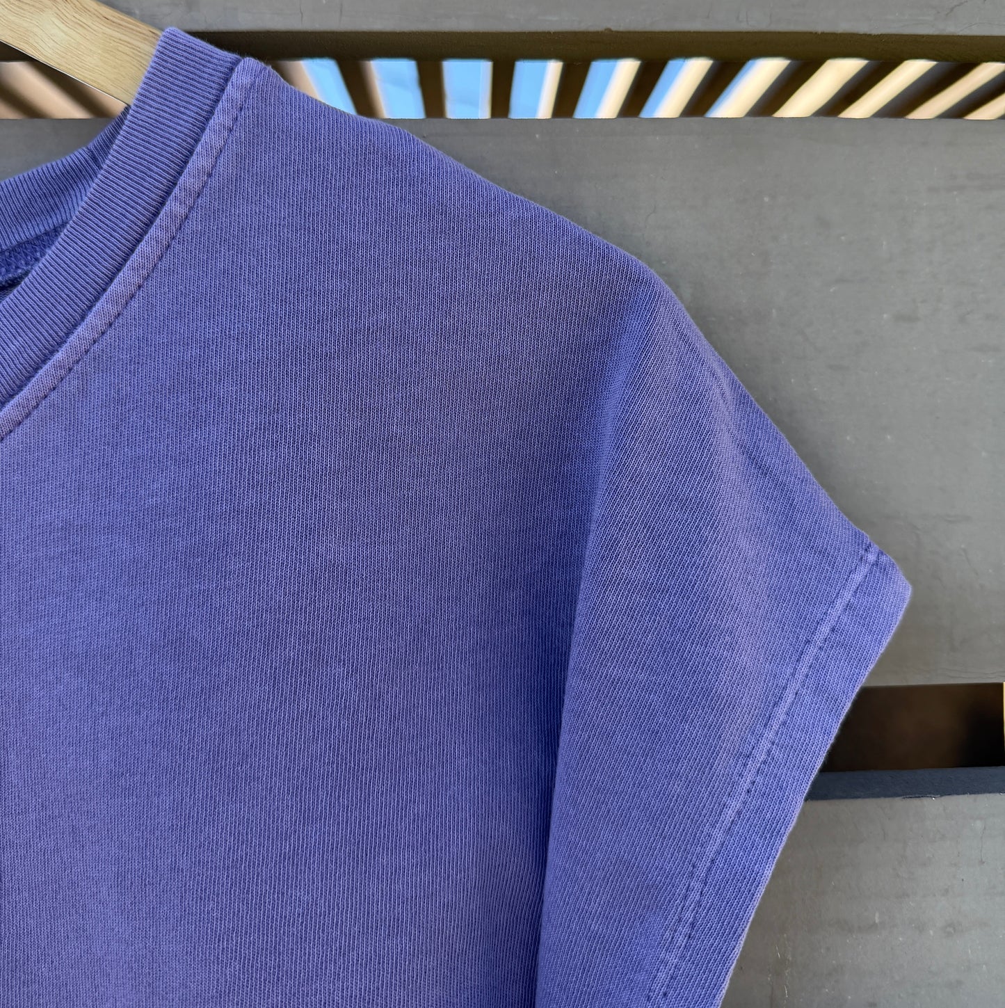 A close-up of a lavender-purple short-sleeve t-shirt hanging on a wooden hanger against a gray concrete wall with a slatted wooden beam.