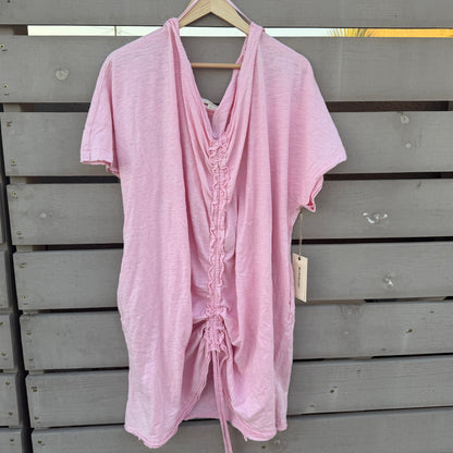 A light pink short-sleeve linen tunic with a gathered front and drawstring detail, hanging on a wooden hanger against a gray wooden slat fence.