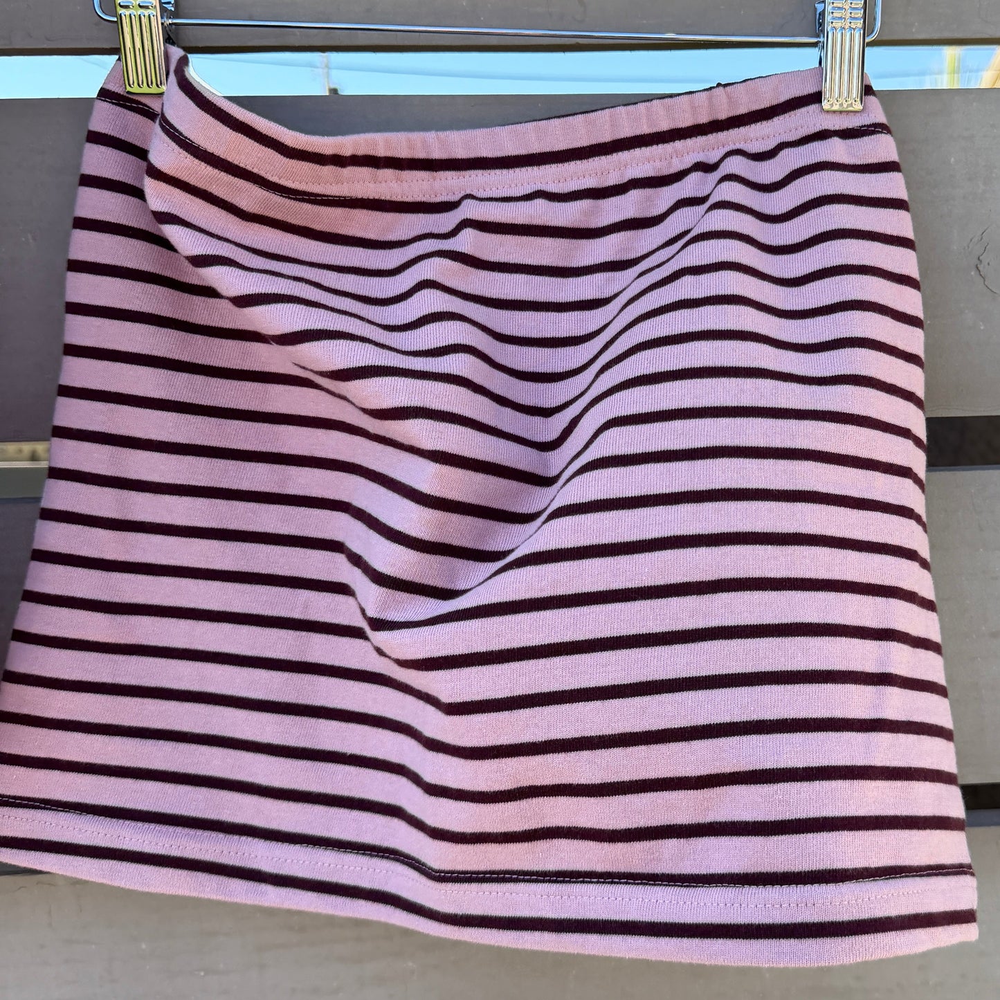 A pink and dark red striped tube top hanging from a wooden clothes hanger against a gray wooden slat wall.